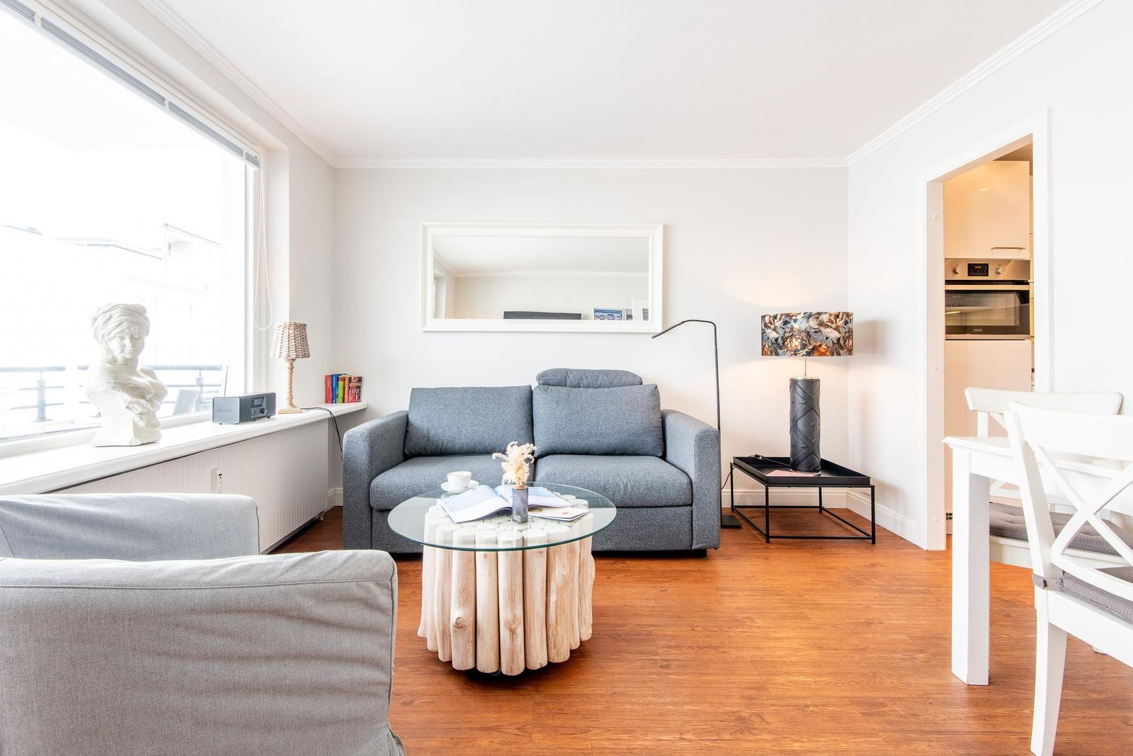 Wohnzimmer mit grauem Sofa, Glas-Tisch und Holzboden. Küche im Hintergrund sichtbar.