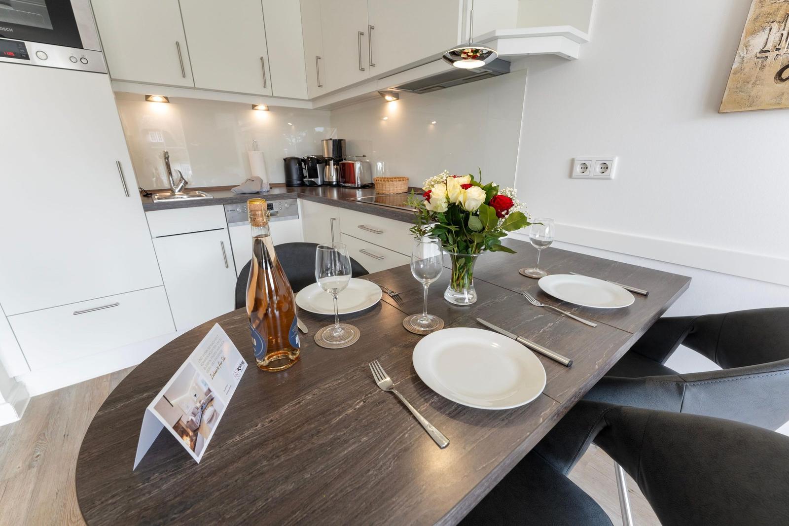 Dining area with table, chairs, and flower arrangement. Kitchen with white cabinets and worktop.