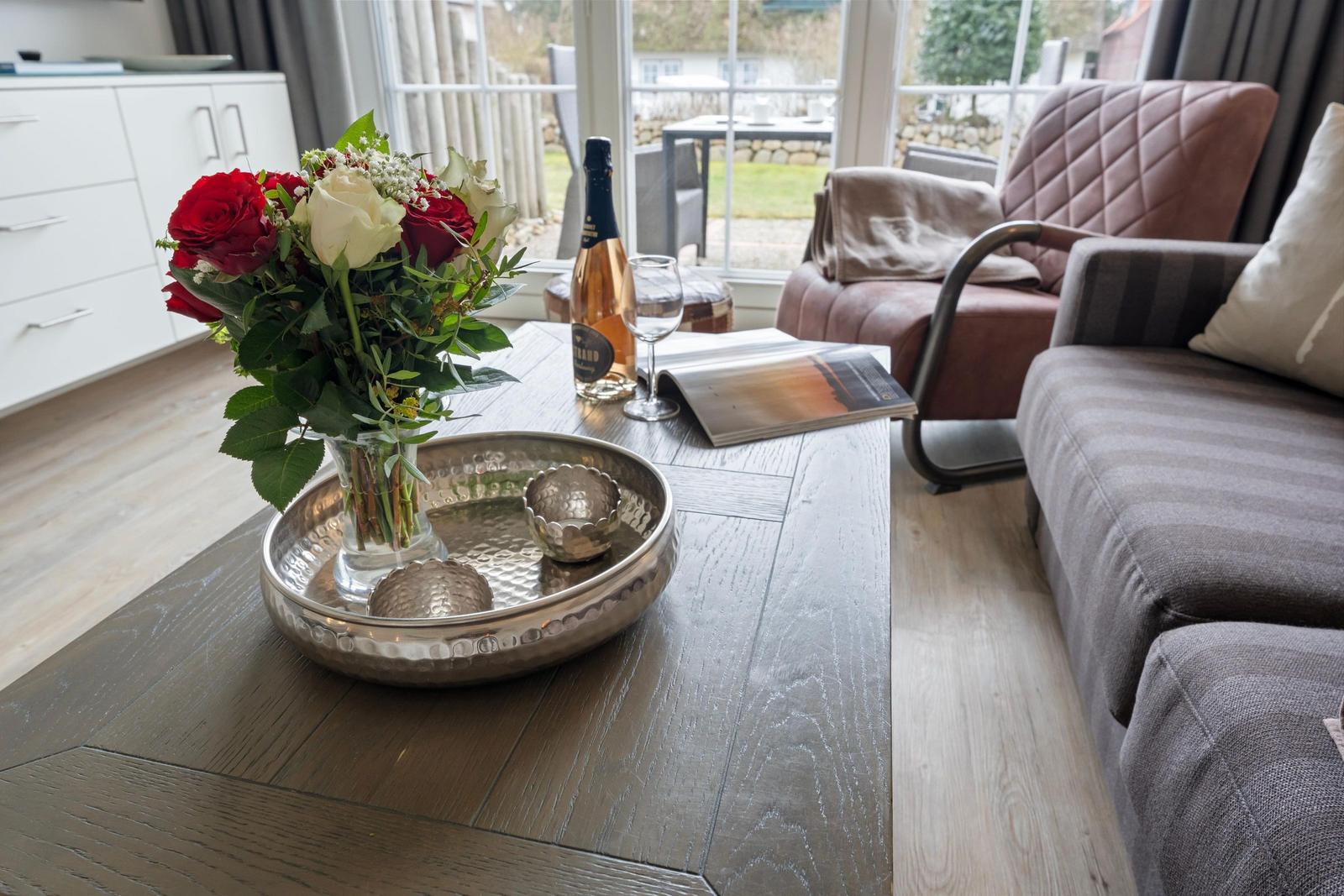 Wooden table with flowers, wine, and magazine in living room with sofa and armchair.