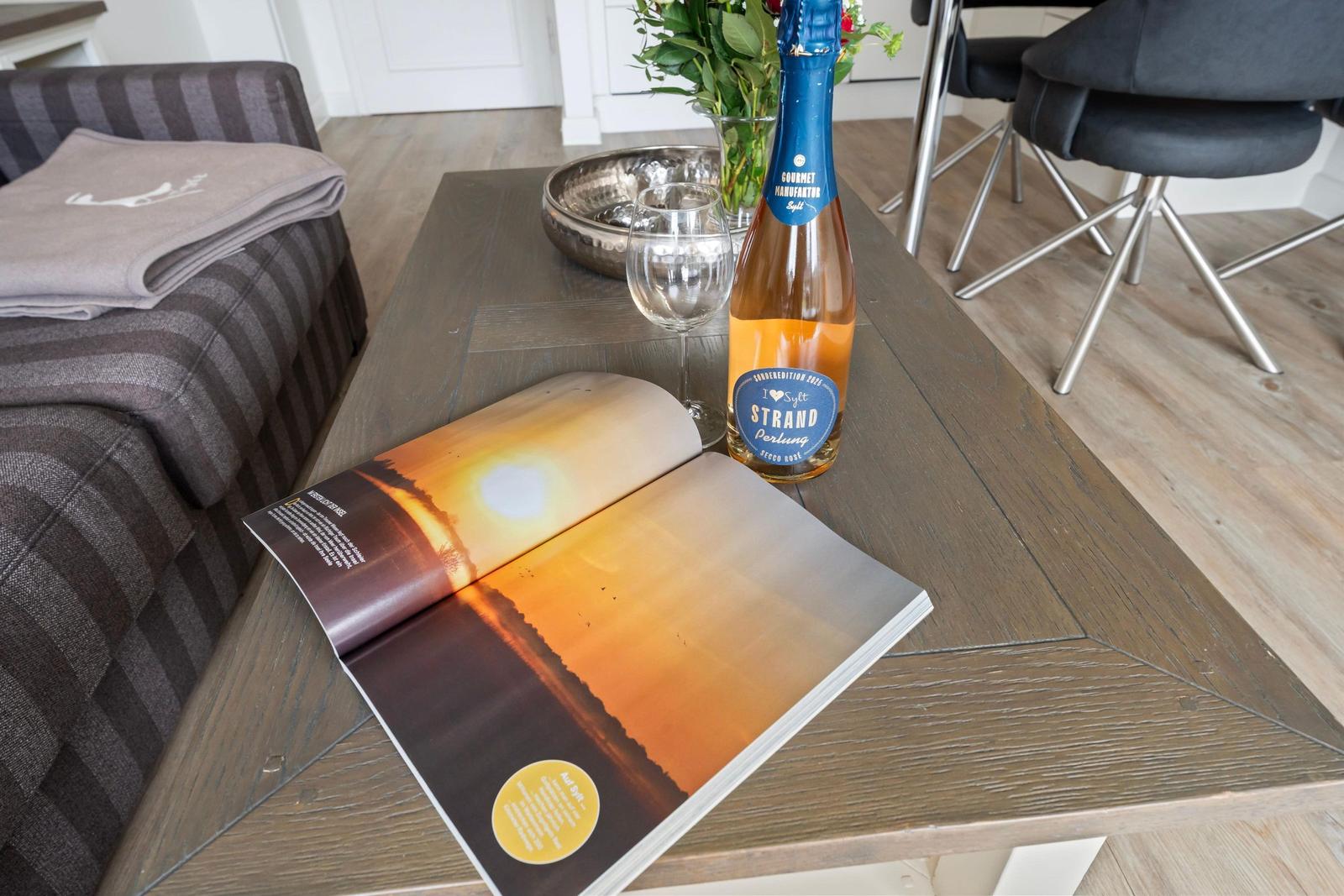 On the table is a bottle of sparkling wine, a glass, and an open magazine.