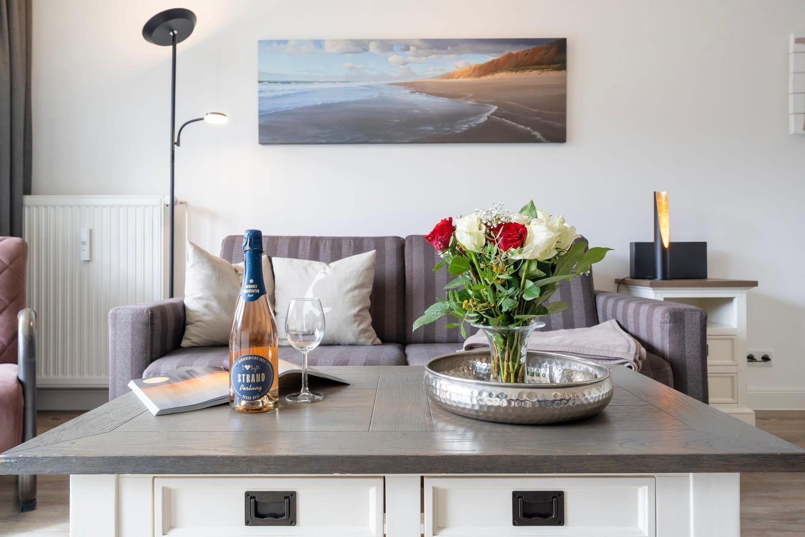 Living room with sofa, table, flowers, and bottle. Wall art shows beach.