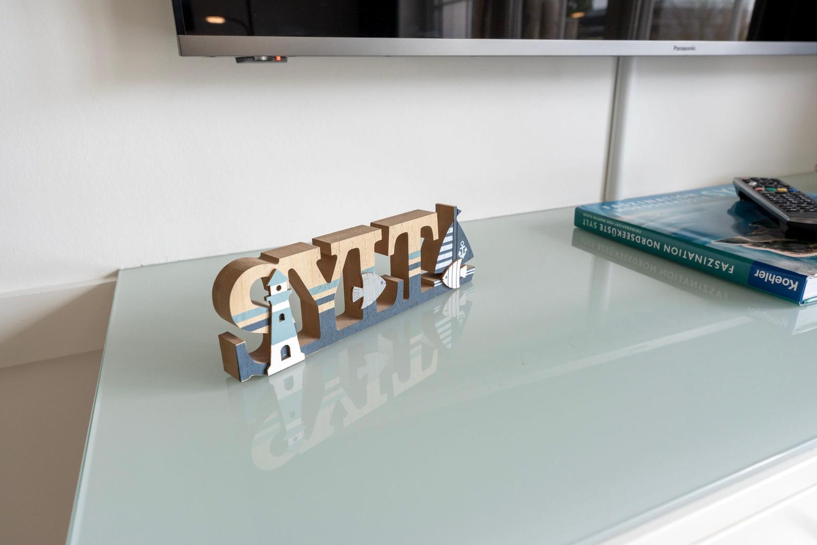 On the table, a wooden object with the word Sylt and maritime motifs stands. Beside it, a book and a remote control lie.