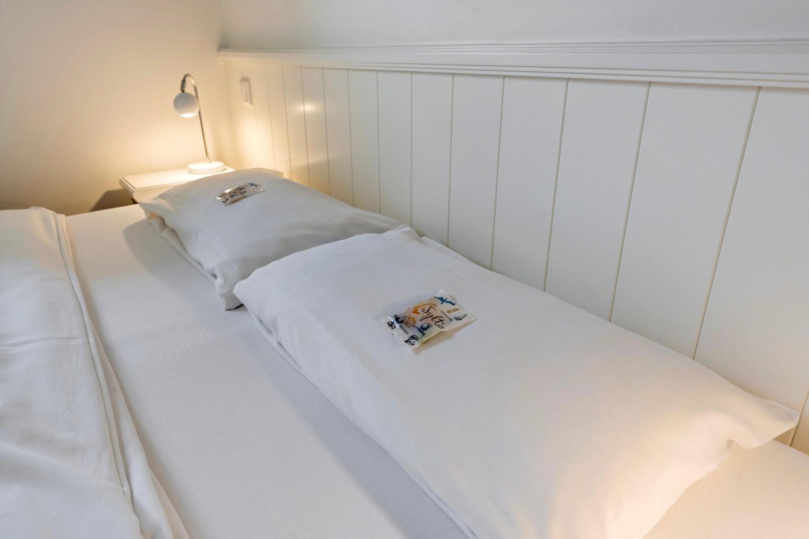 A bed with white bedding and two pillows. A bedside lamp is mounted on the wall.