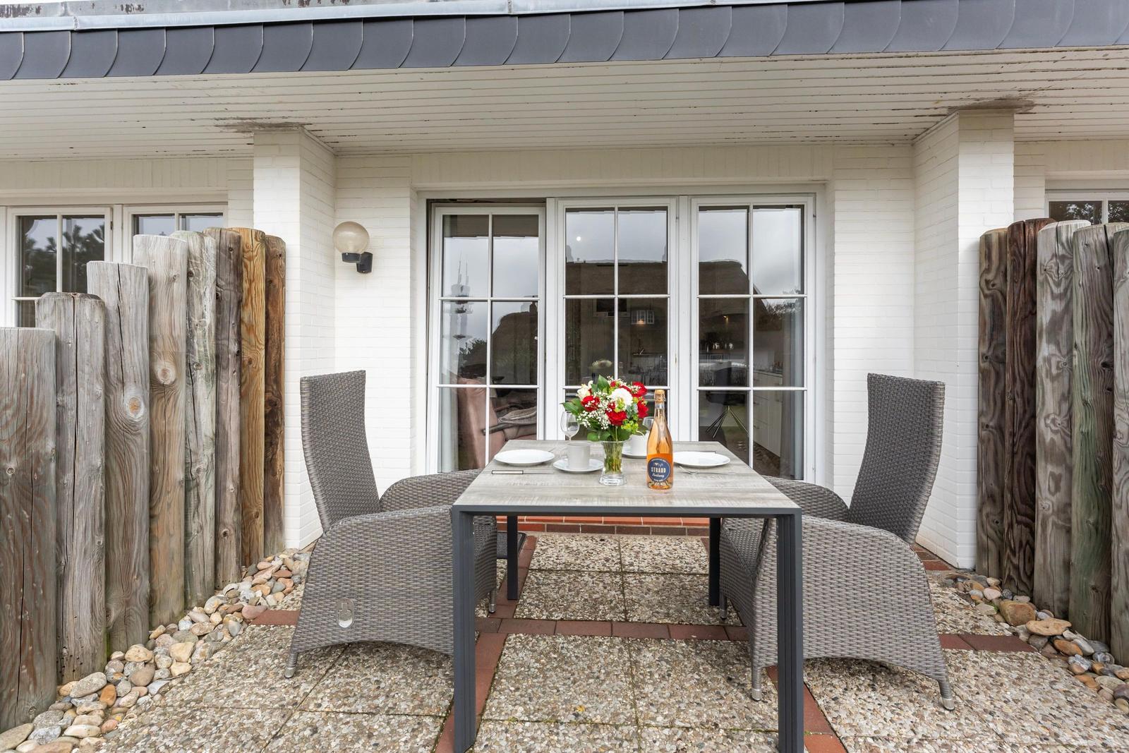 Terrace with table, chairs, and flowers in front of white house wall.