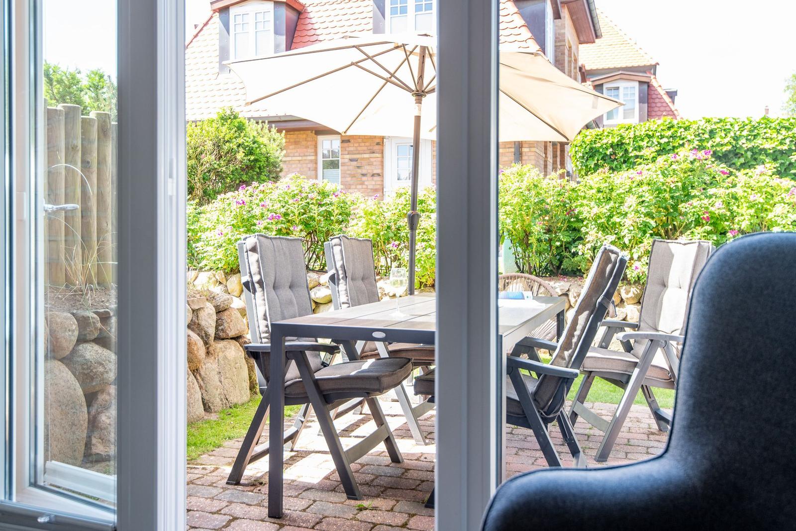 Terrasse mit Tisch, Stühlen und Sonnenschirmen. Blick auf Garten mit Sträuchern und Steinmauer.