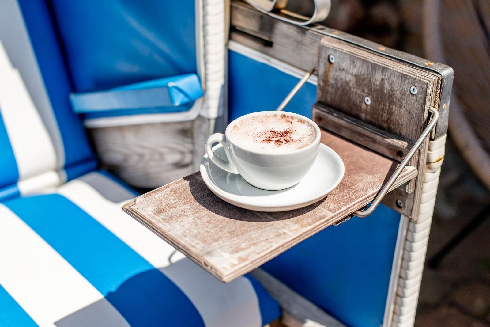 Eine Tasse Cappuccino mit Kakaopulver steht auf einem Holztisch neben einem blauen Liegestuhl.