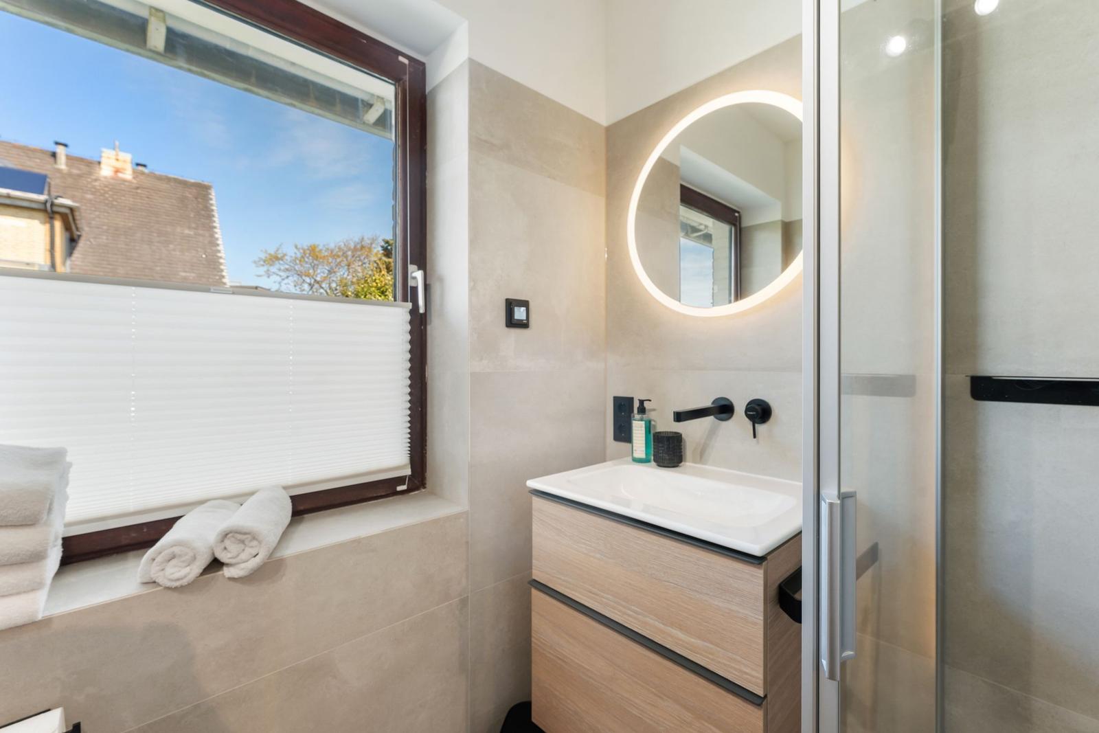 Badezimmer mit Waschbecken, Dusche und Fenster mit Blick auf Dach und Baum.