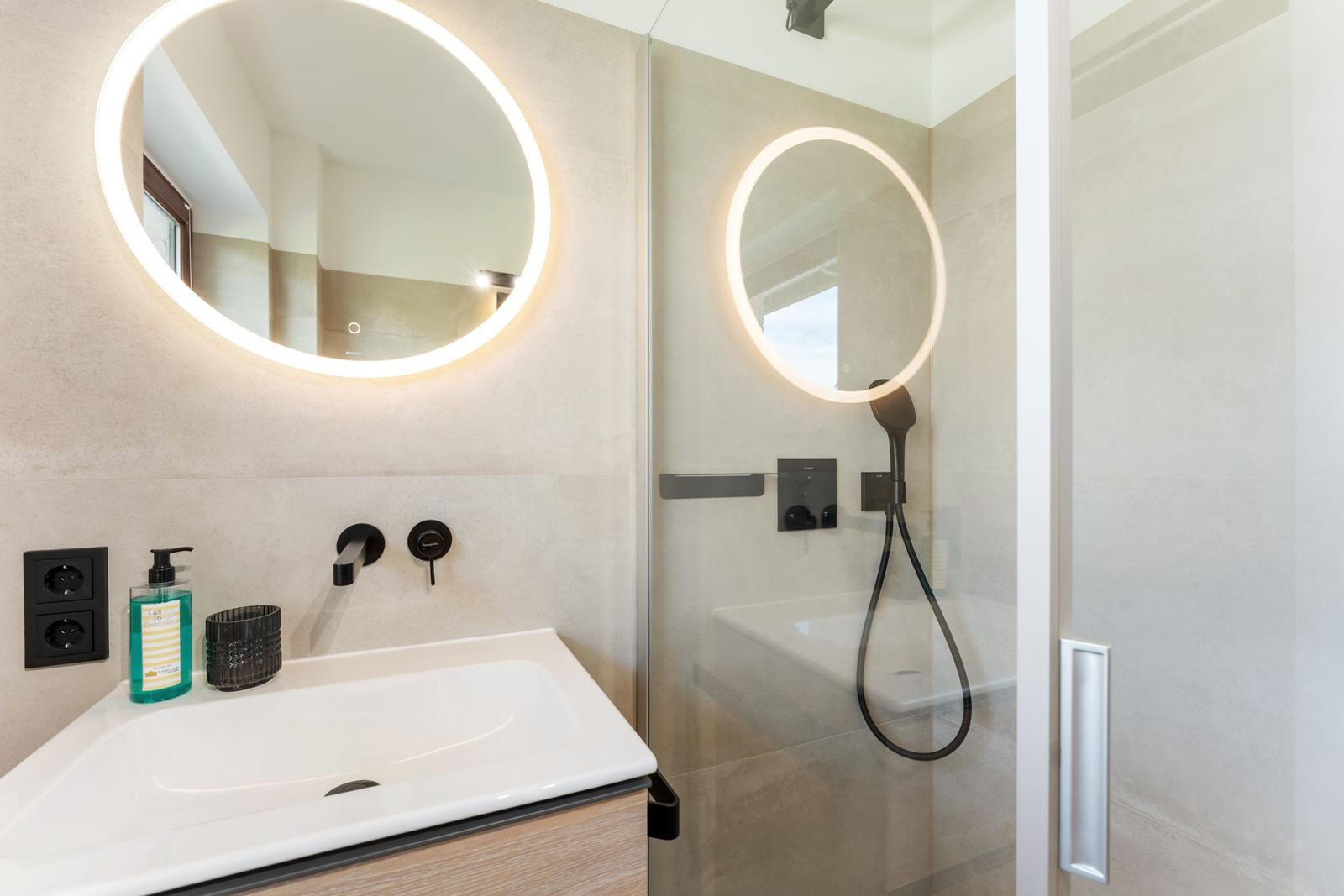Moderne Dusche mit Glaswand und runden beleuchteten Spiegel. Waschbecken mit schwarzen Armaturen.