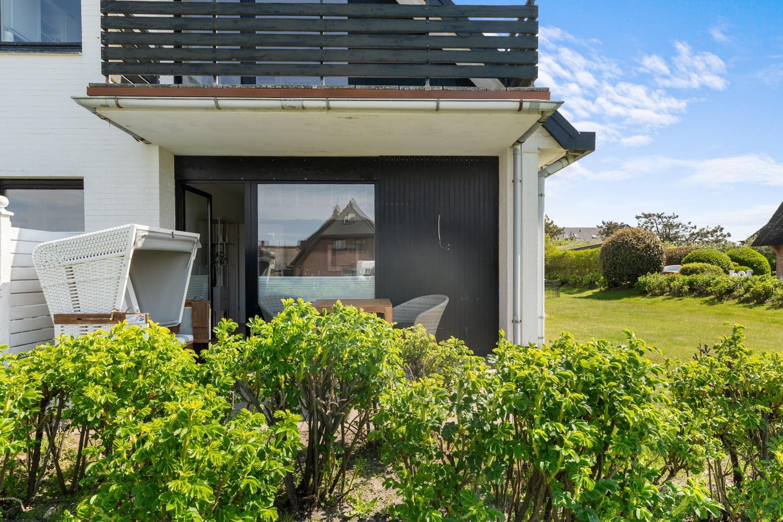 Wohnung mit Balkon, Garten und Terrasse. Weiße Hauswand, grüne Hecken und Sitzmöbel.