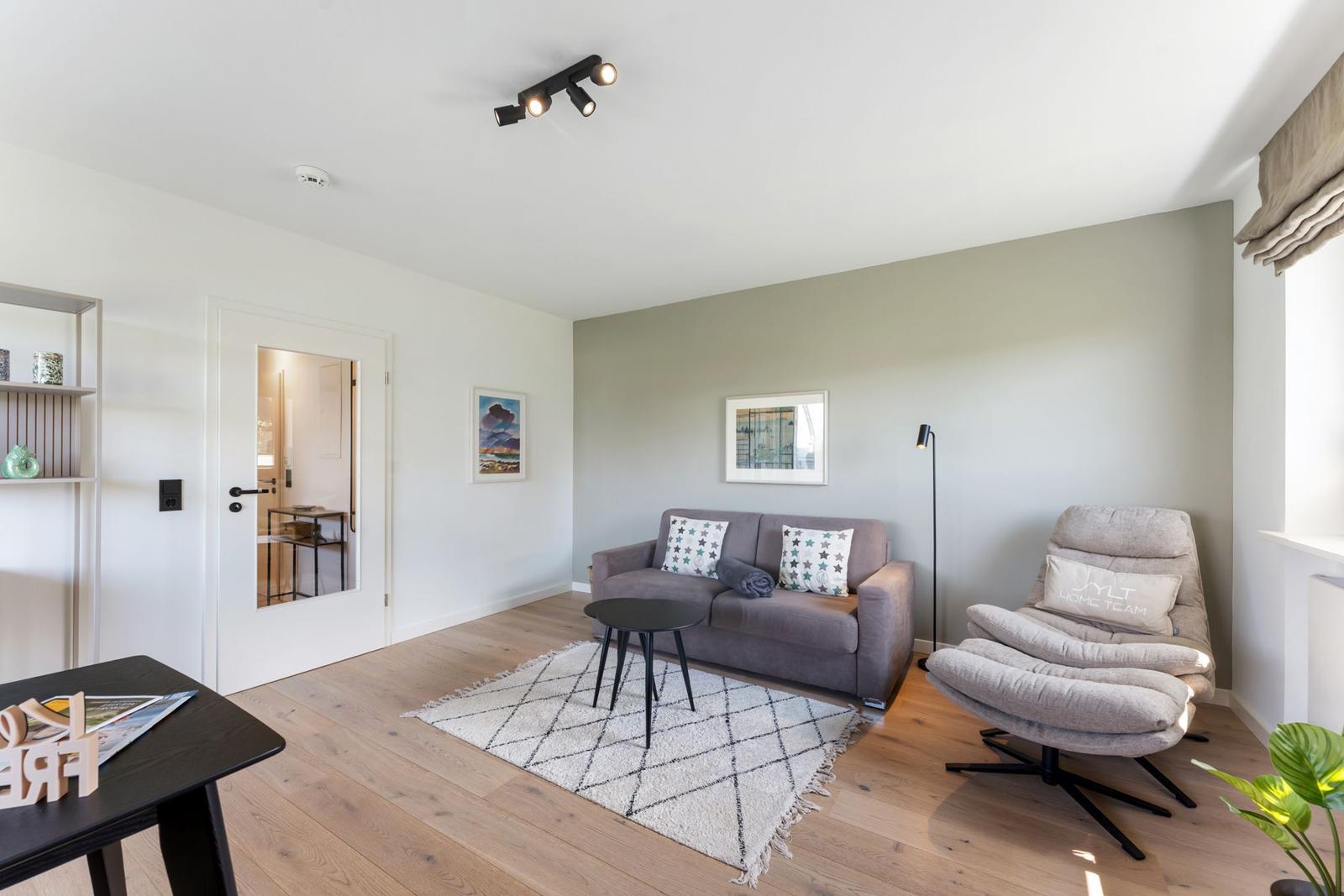 Wohnzimmer mit Sofa, Sessel, Teppich und Holzboden. An der Wand hängen zwei Bilder.