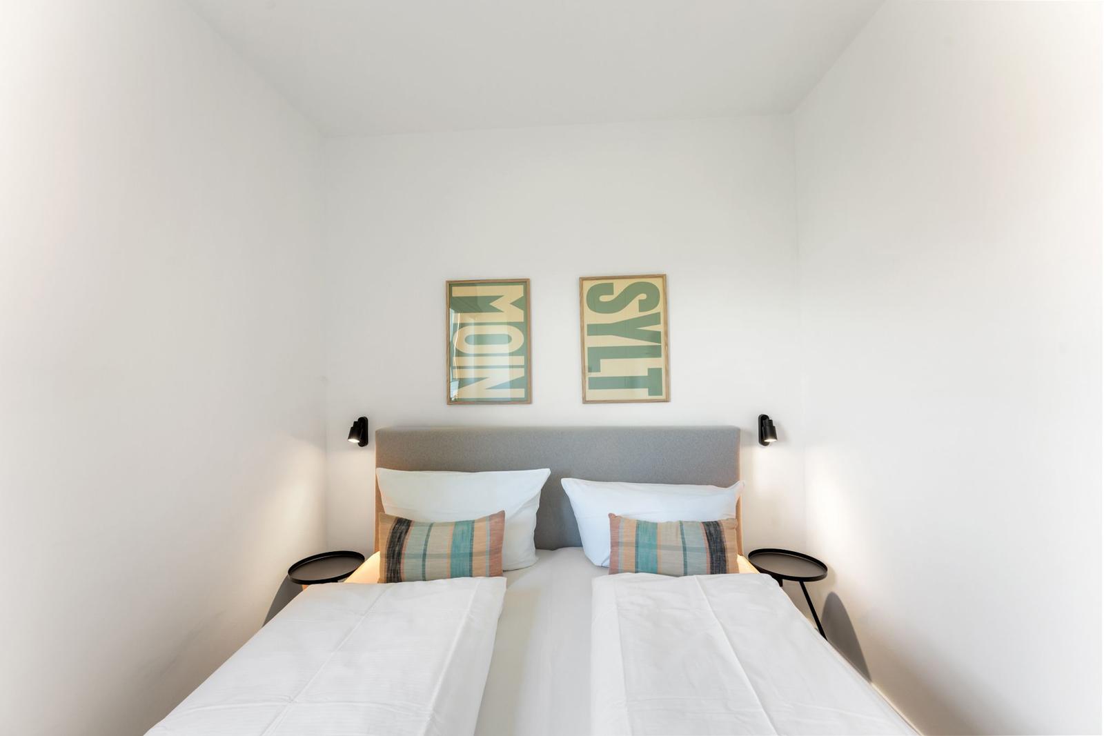 Schlafzimmer mit Doppelbett, zwei Wandlampen und zwei Bildern mit Text.