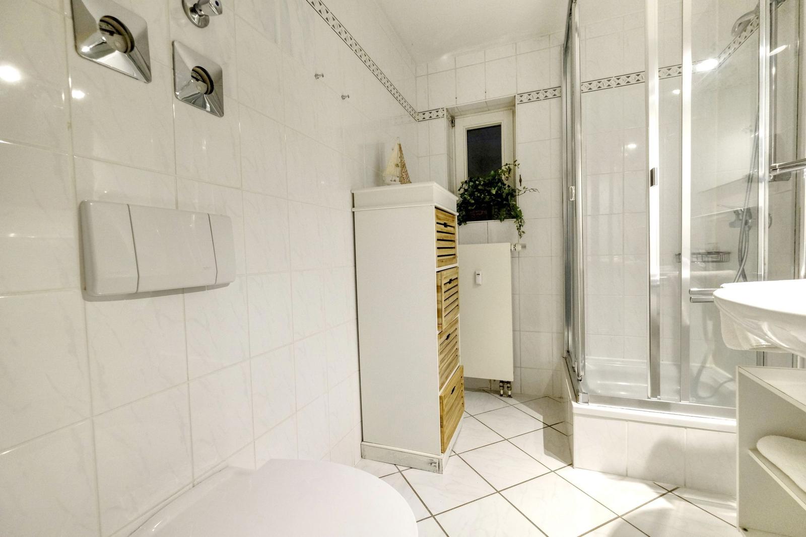 Badezimmer mit Dusche, Waschbecken und Toilette. Weiße Fliesen und Holzschrank.