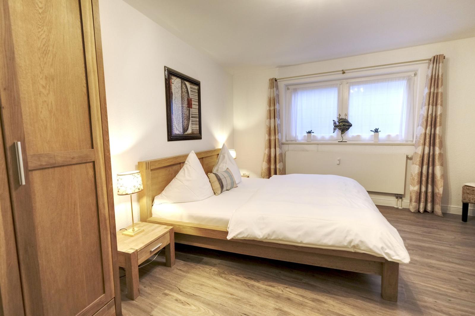 Schlafzimmer mit Bett, Nachttisch, Lampe und Fenster mit Vorhängen.