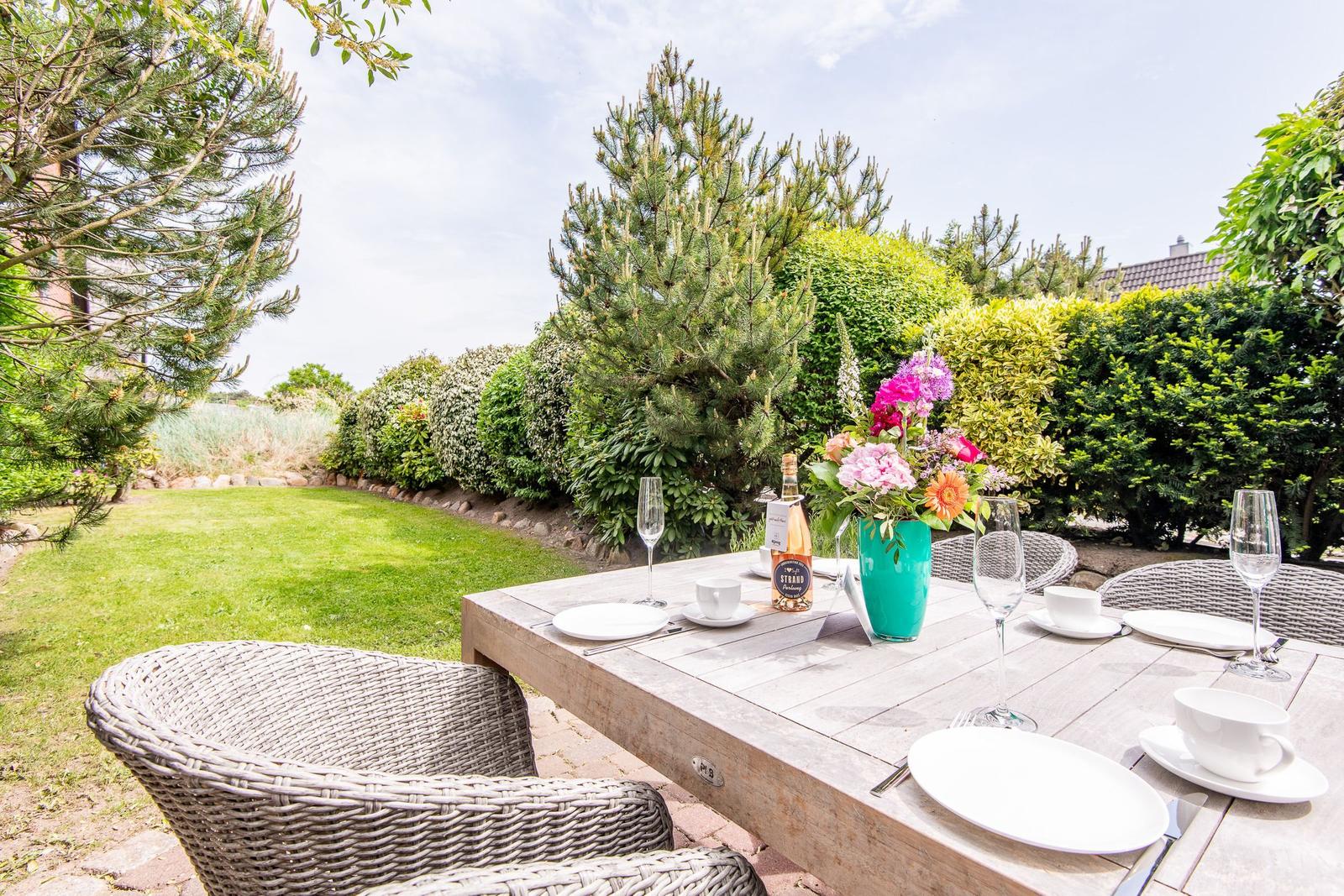 Garten-Terrasse mit gedecktem Tisch, Blumen und Stühlen. Grünflächen und Bäume im Hintergrund.