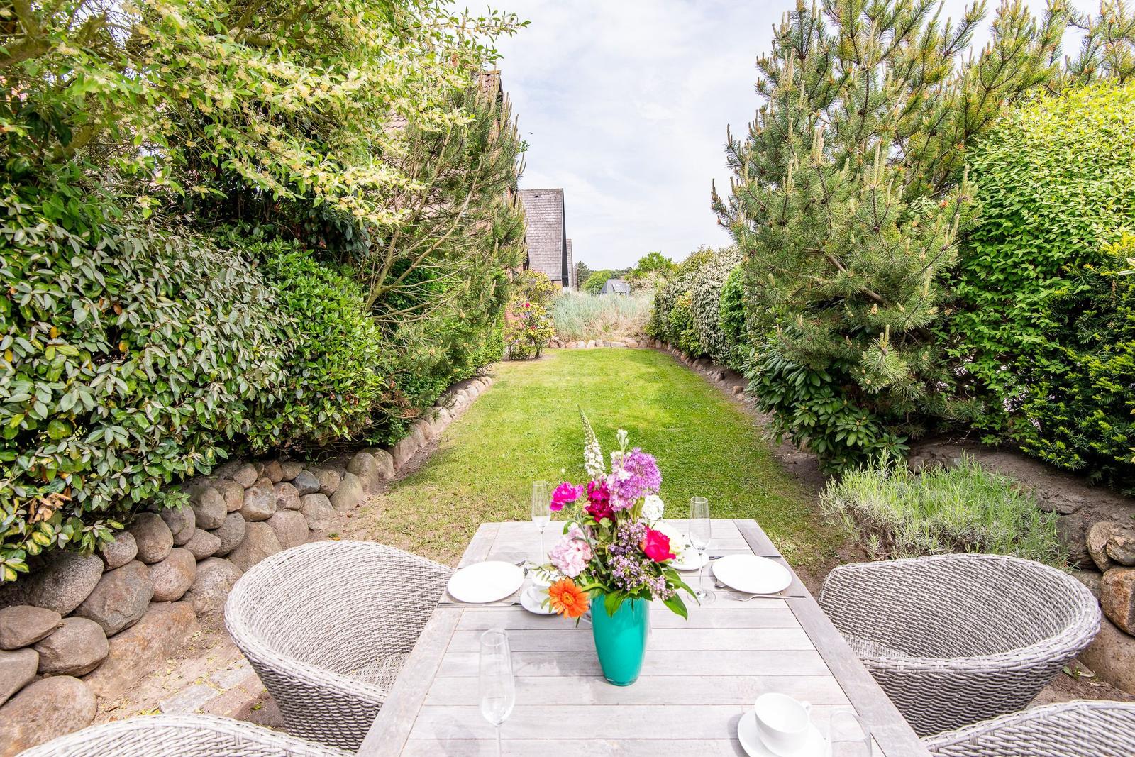 Garten mit Tisch, Stühlen und Blumenarrangement zwischen Hecken und Bäumen.