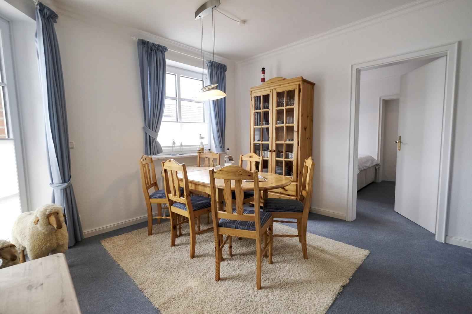 Dining room with wooden table, chairs, and sideboard. Windows with curtains. View into bedroom.