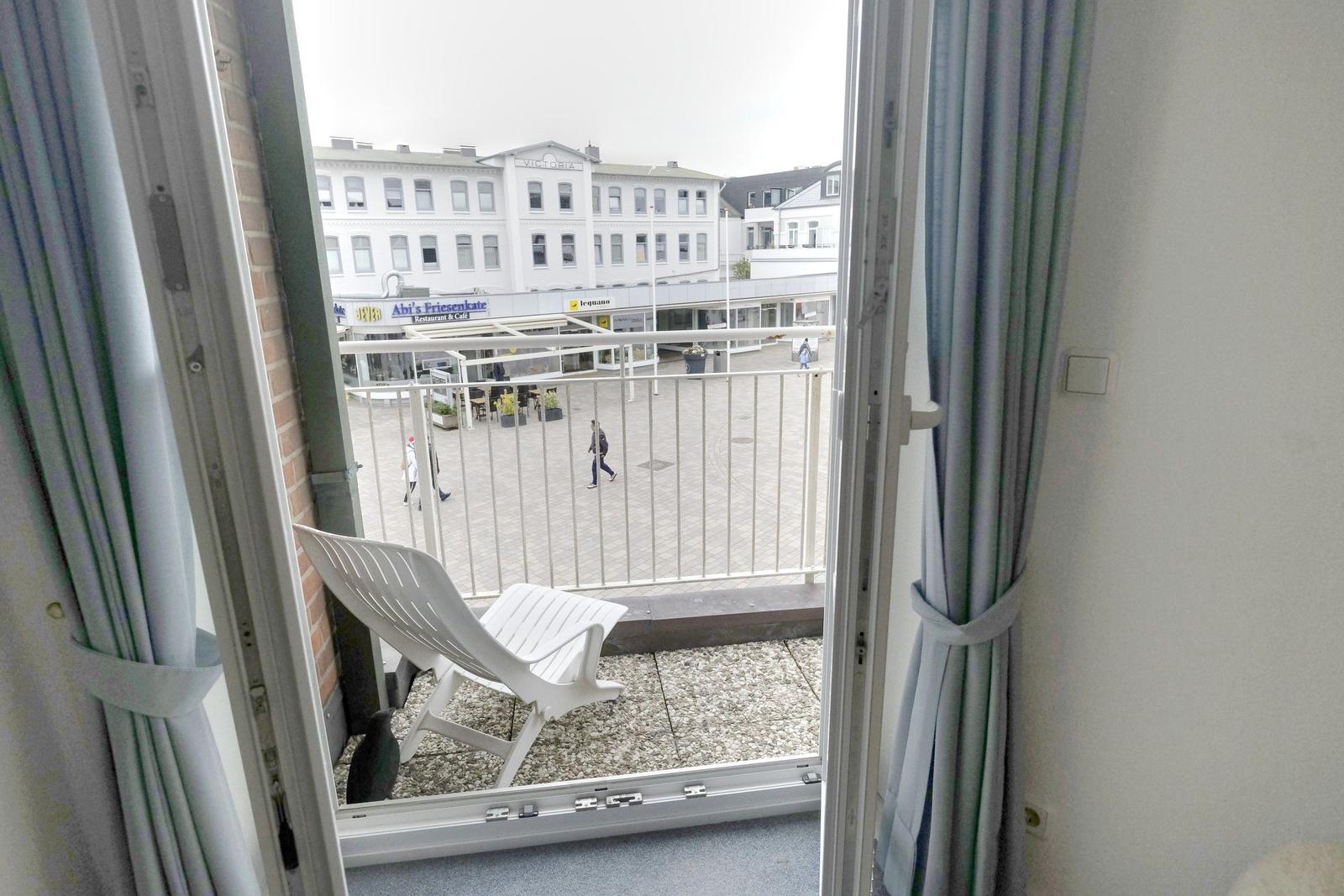 Balcony with white lounge chair and view of busy square with shops and pedestrians.