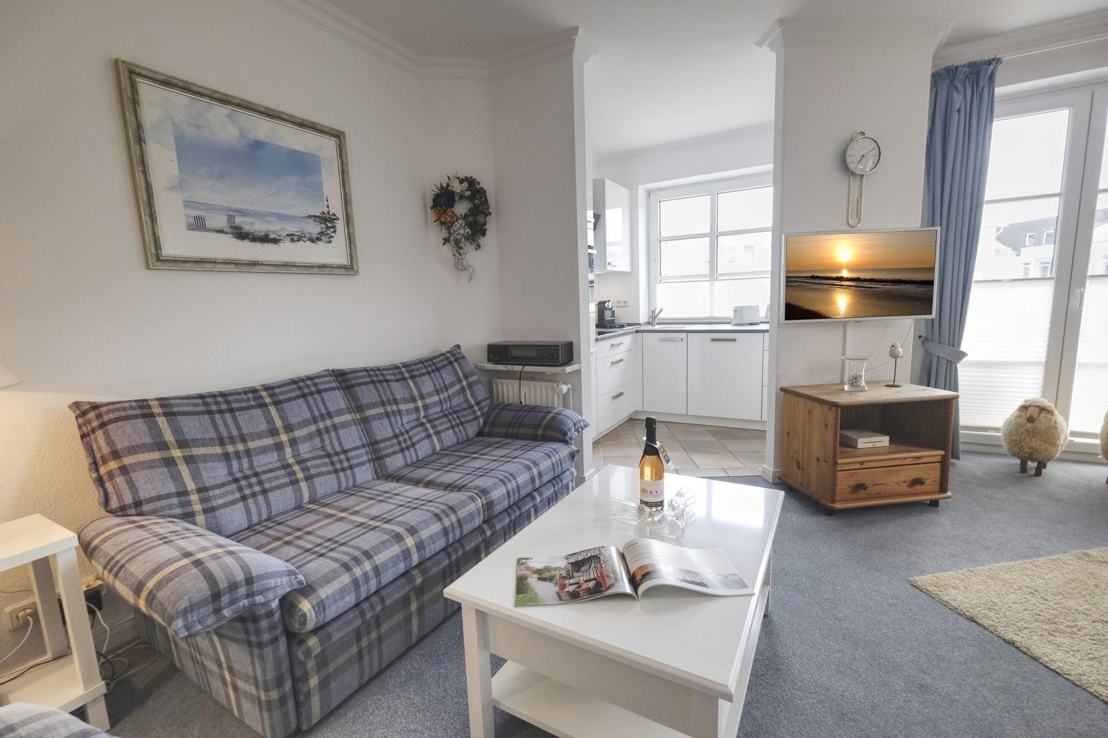 Living room with sofa, coffee table, and TV. Kitchen in background. Windows with curtains.