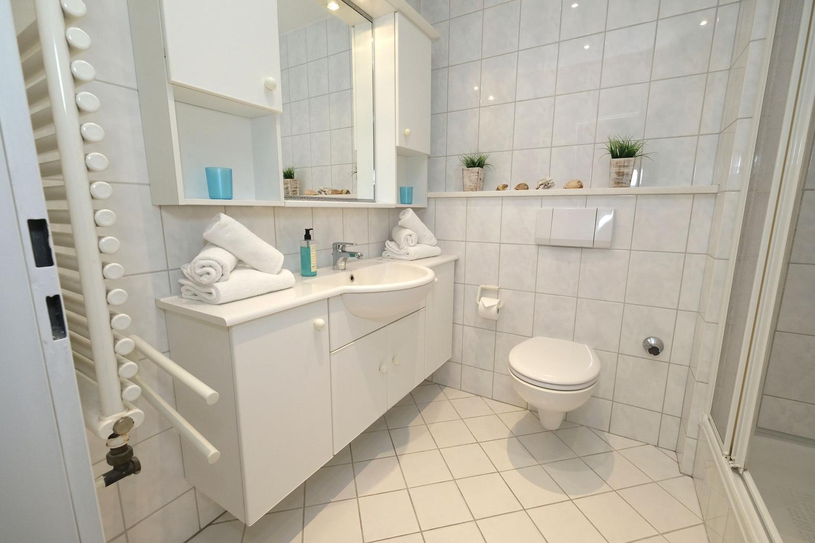 Badezimmer mit Waschbecken, Toilette und Dusche. Weiße Fliesen und Handtücher.