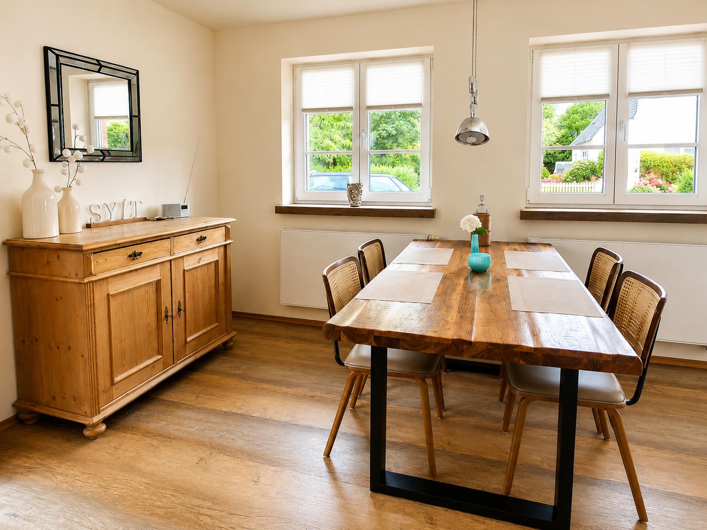 Esszimmer mit Holztisch, Stühlen und Fenstern. An der Wand steht ein Sideboard mit Spiegel.