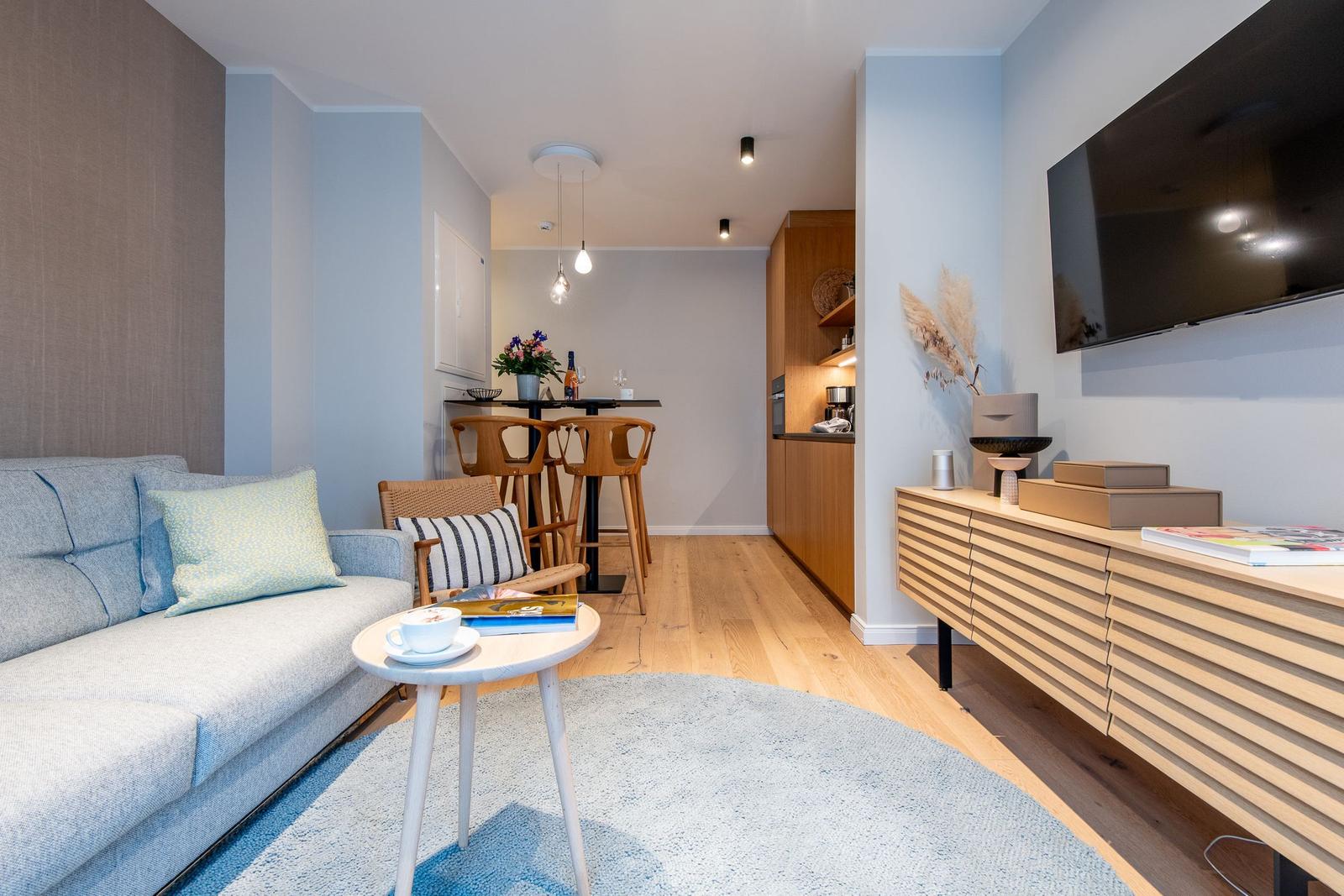 Living area with sofa, table, and TV. Kitchen and seating area in the background.