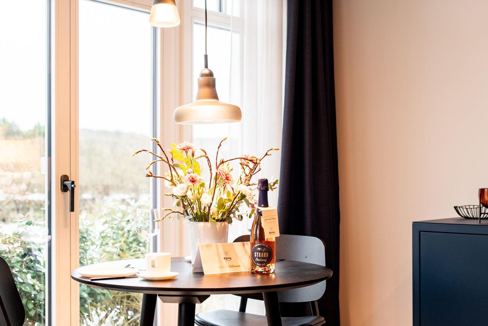Runder Tisch mit Blumen, Flasche und Tasse vor Fenster mit Blick auf Garten.