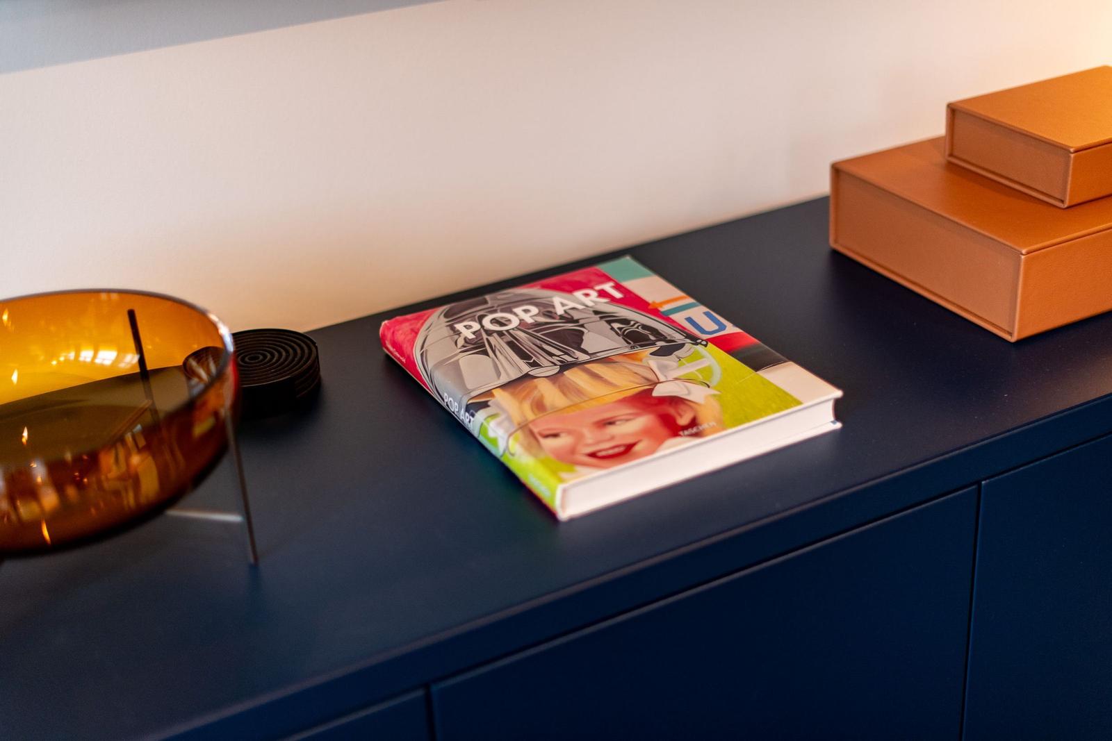 Ein Buch über Pop Art liegt auf einem dunkelblauen Schrank neben einer braunen Schachtel und einer Glaslaterne.