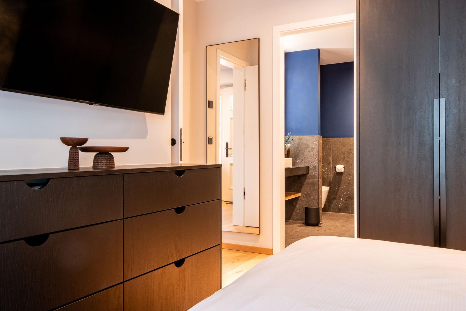 Schlafzimmer mit Bett, Kleiderschrank und Fernseher. Badezimmer mit blauer Wand sichtbar.