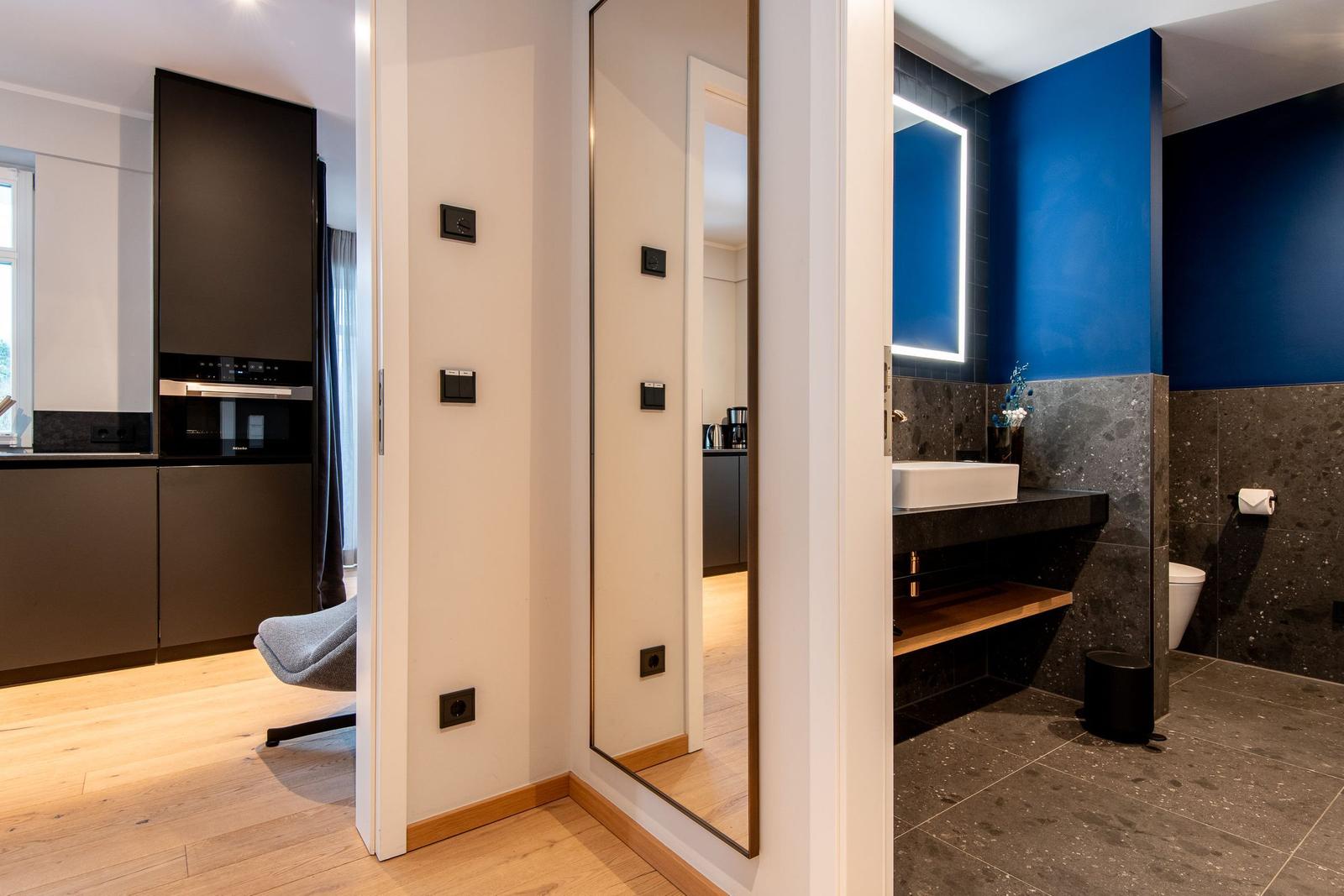 Badezimmer mit blauer Wand und Waschtisch. Küche mit dunklen Schränken sichtbar.