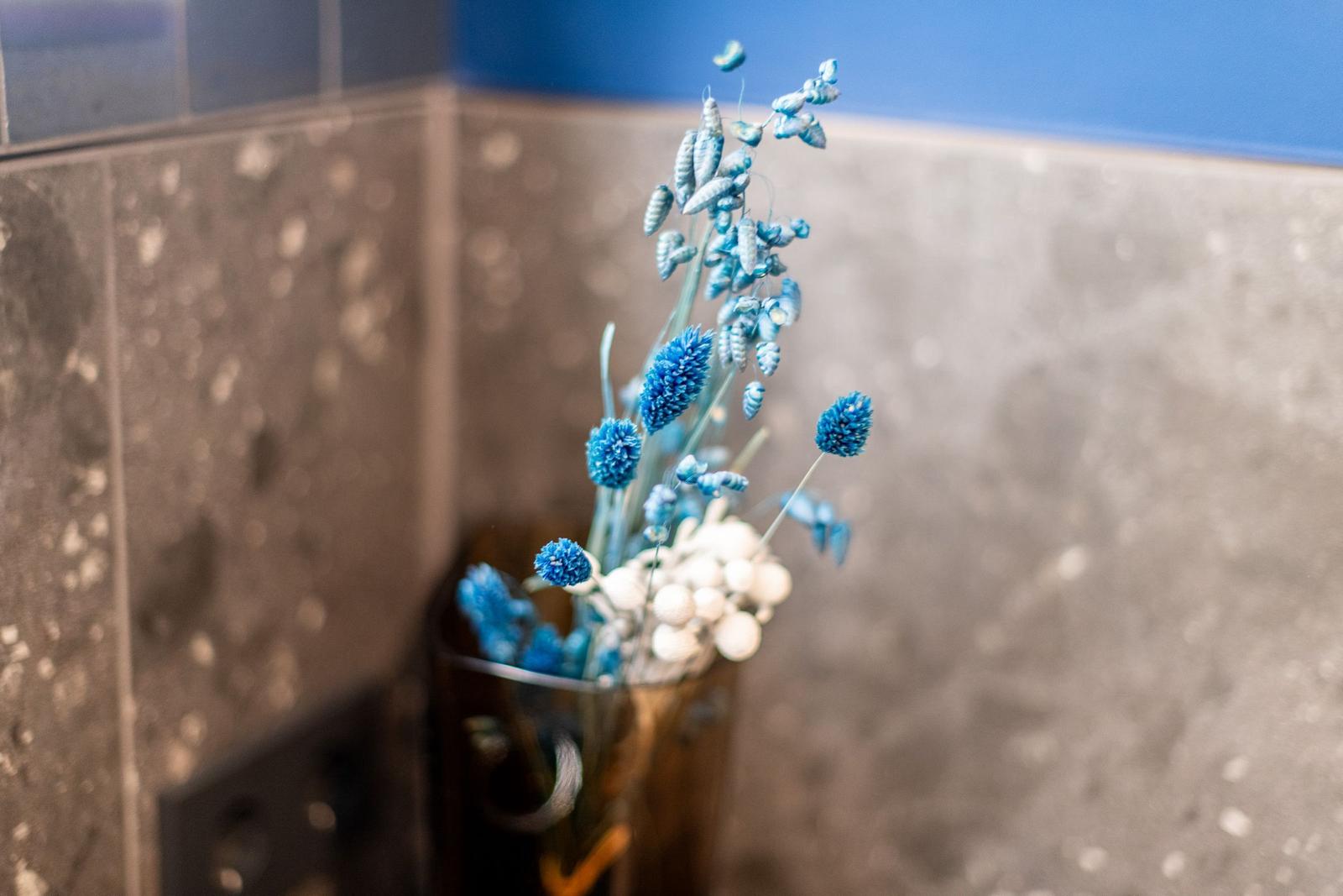 Blauer Blumenschmuck in einem Glas auf einer Wand mit grauen Fliesen.
