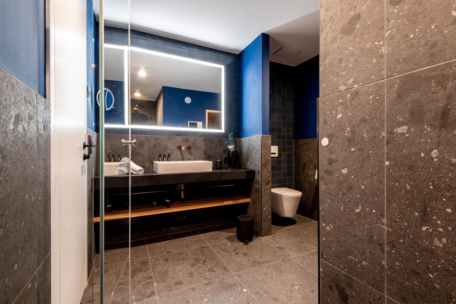 Badezimmer mit Dusche, Waschbecken und Toilette. Dunkle Fliesen und blauer Akzent.