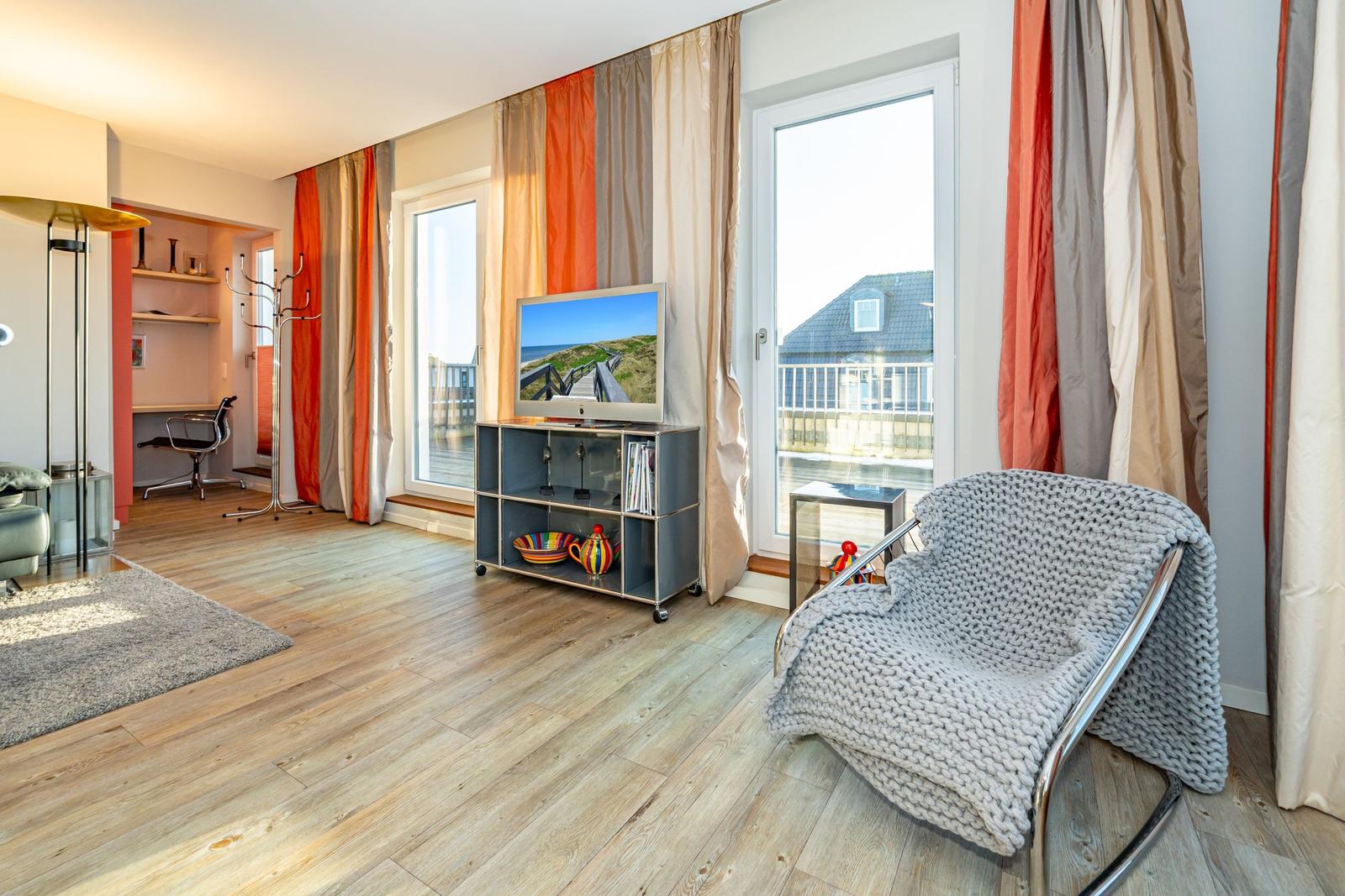 Wohnzimmer mit TV, Sessel und Blick auf Balkon. Holzboden und farbige Vorhänge.