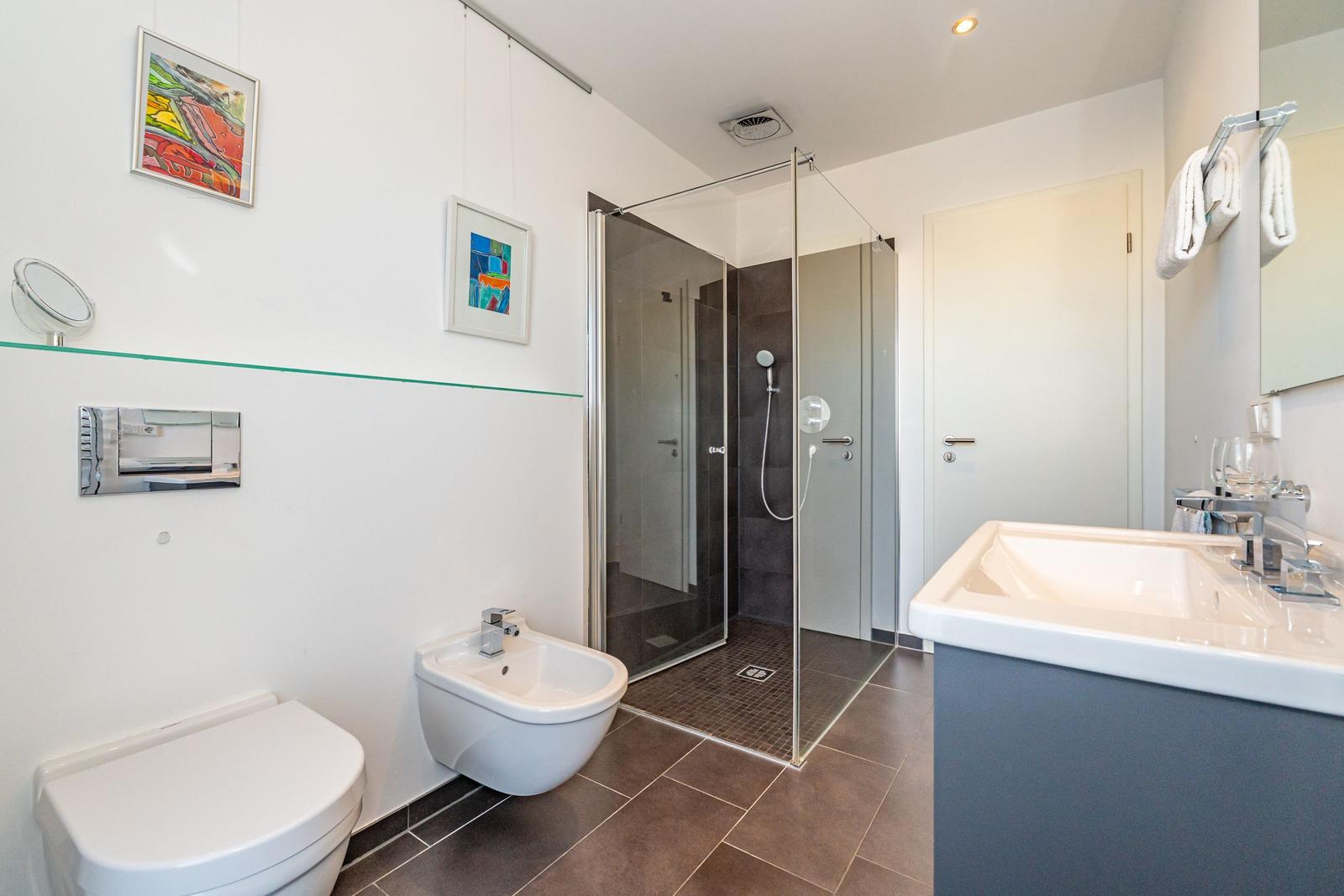Badezimmer mit Dusche, Waschbecken und Toilette. Wandbilder und Handtücher sichtbar.