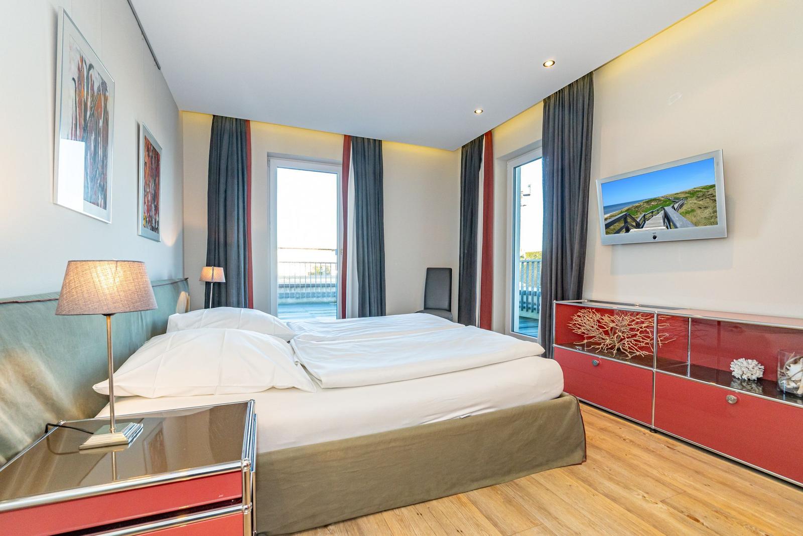 Schlafzimmer mit Bett, Fenstern, TV und rotem Schrank.