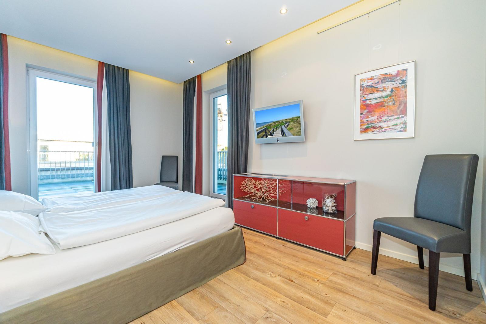 Schlafzimmer mit Doppelbett, zwei Fenstern, rotem Schrank und Stuhl.