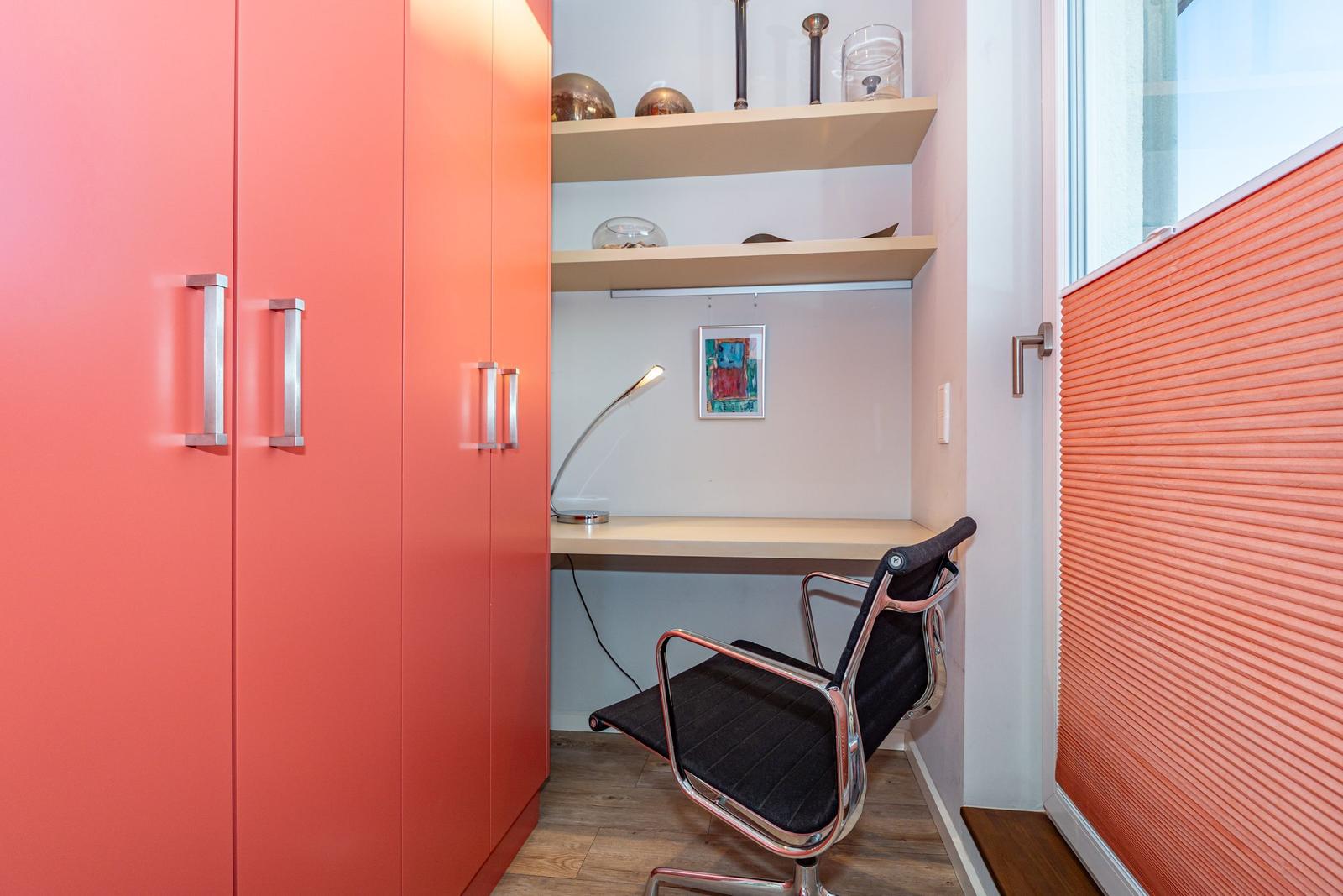 Arbeitsplatz mit Schreibtisch, Stuhl und Regalen. Korallenfarbener Schrank und Wandverkleidung.