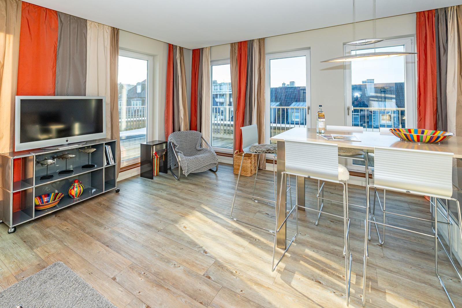 Wohn- und Essbereich mit TV, Tisch und Stühlen, großen Fenstern und Holzboden.