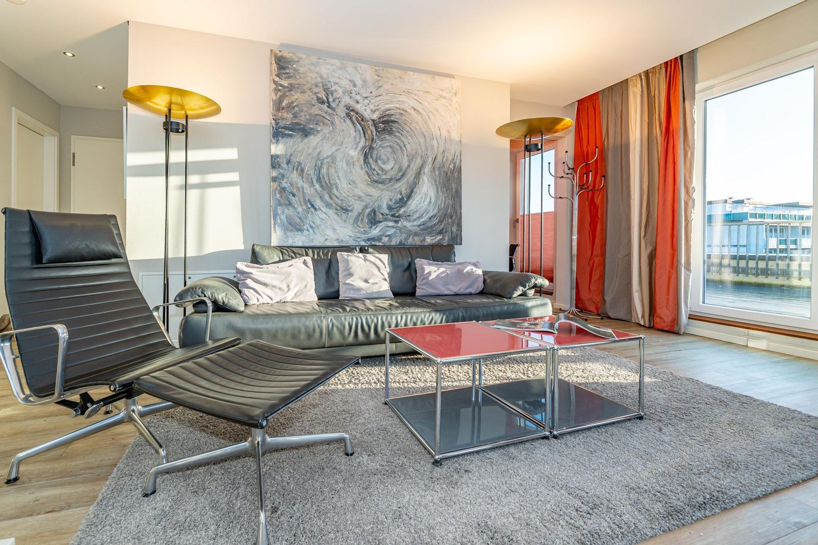 Wohnzimmer mit Sofa, Sessel, Tisch und Wandbild. Große Fenster und Vorhänge.
