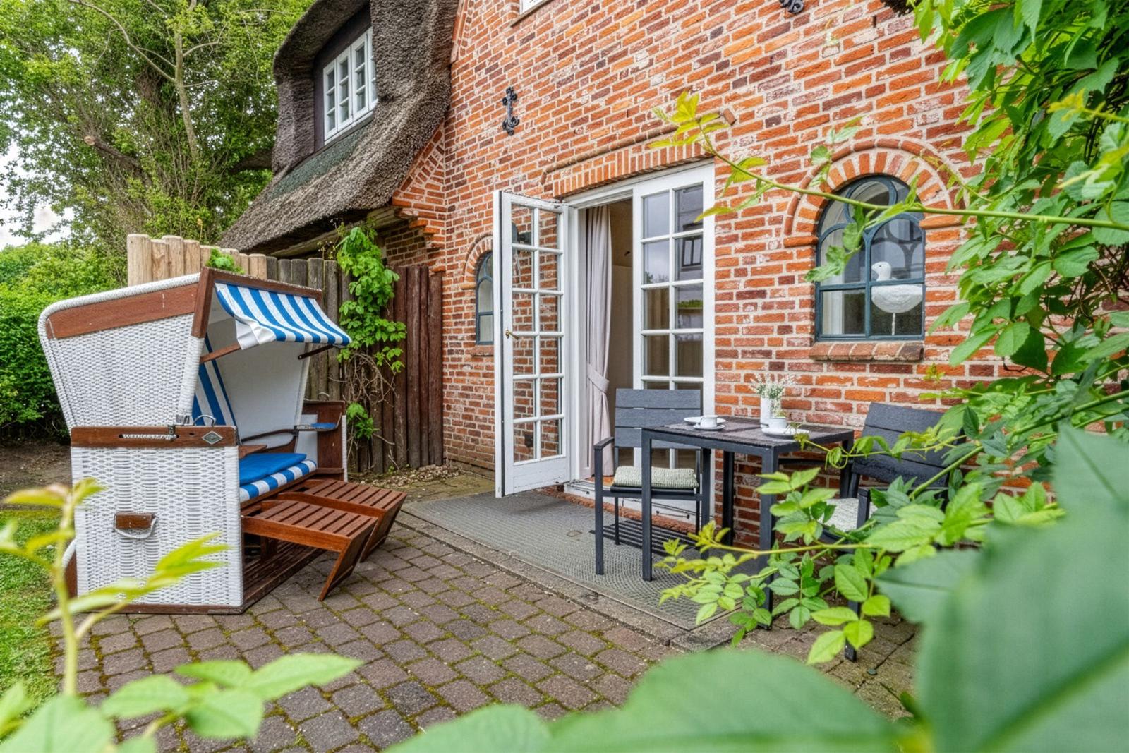 Terrasse mit Gartenliege, Tisch und Stühlen vor rotem Backsteinhaus mit offener Tür.