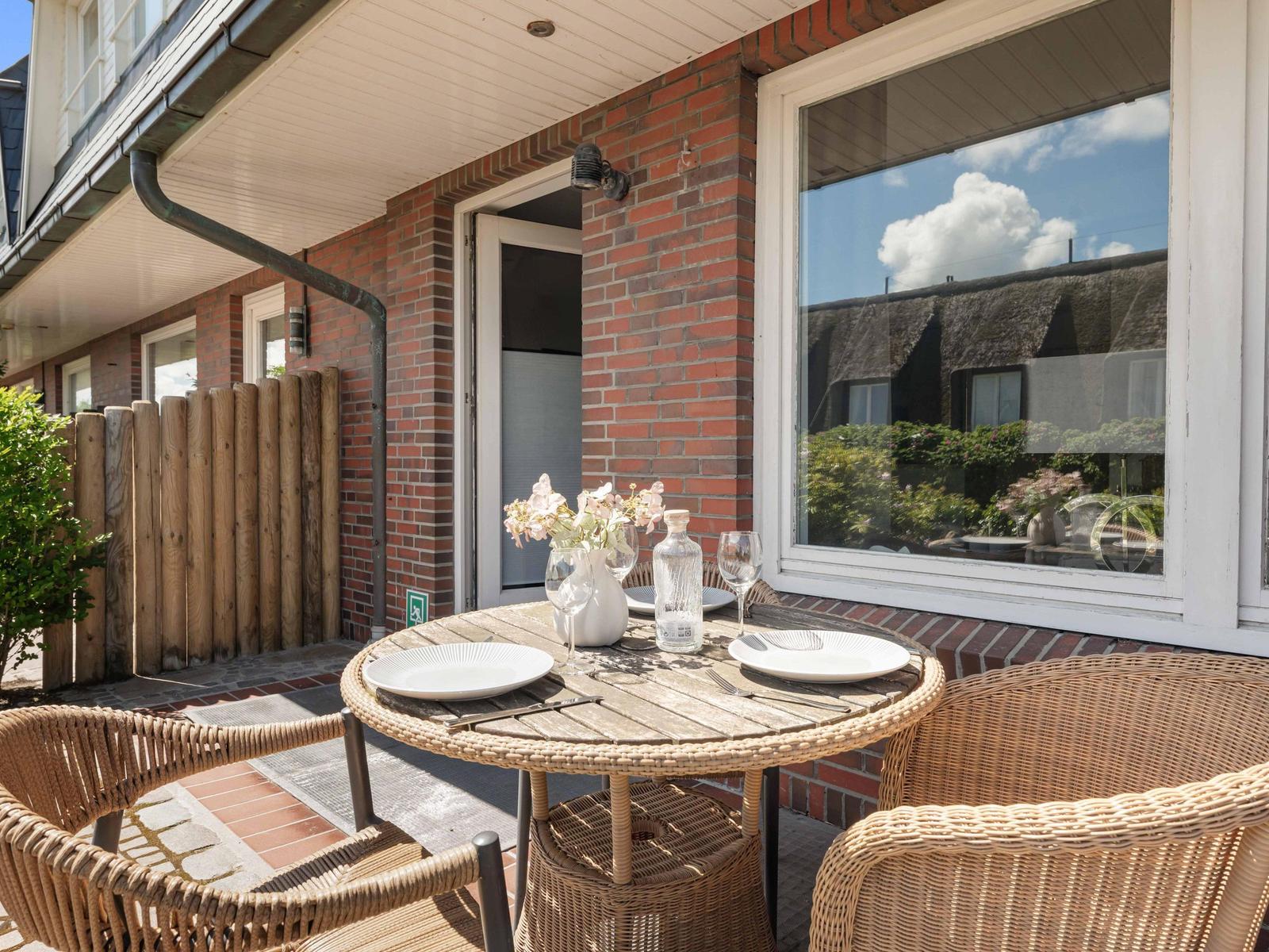 Terrasse mit runden Tisch und Stühlen, Blumen und Geschirr. Blick in Wohnraum mit Fenster.