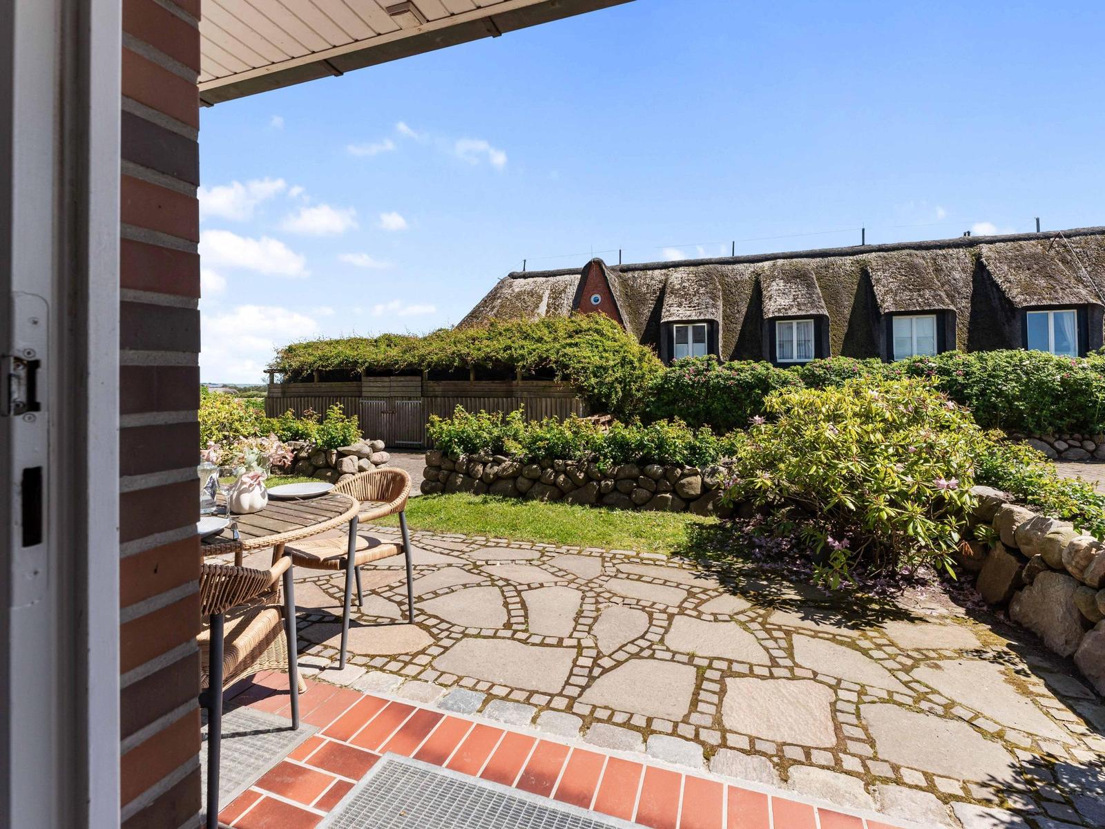 Terrasse mit Tisch und Stühlen, Blick auf traditionelles Haus mit Strohdach und Garten.