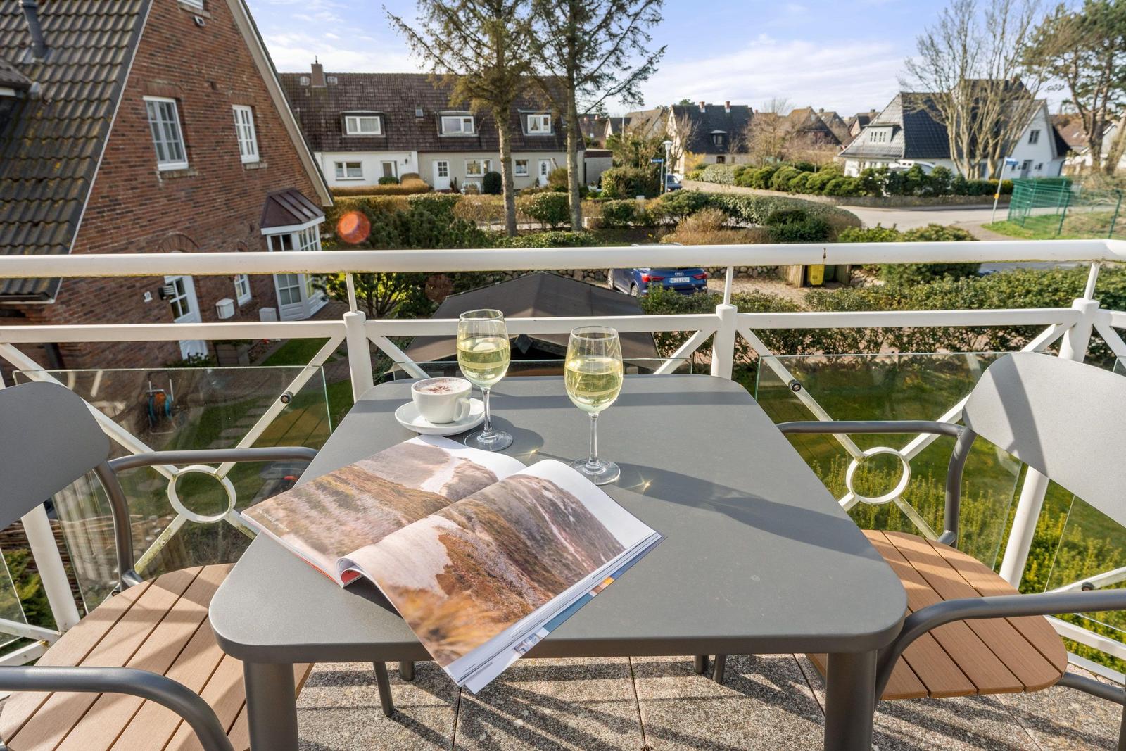 Balkon mit Tisch, zwei Gläser Weißwein, Kaffeetasse und Zeitschrift. Blick auf Wohnhäuser und Grünflächen.