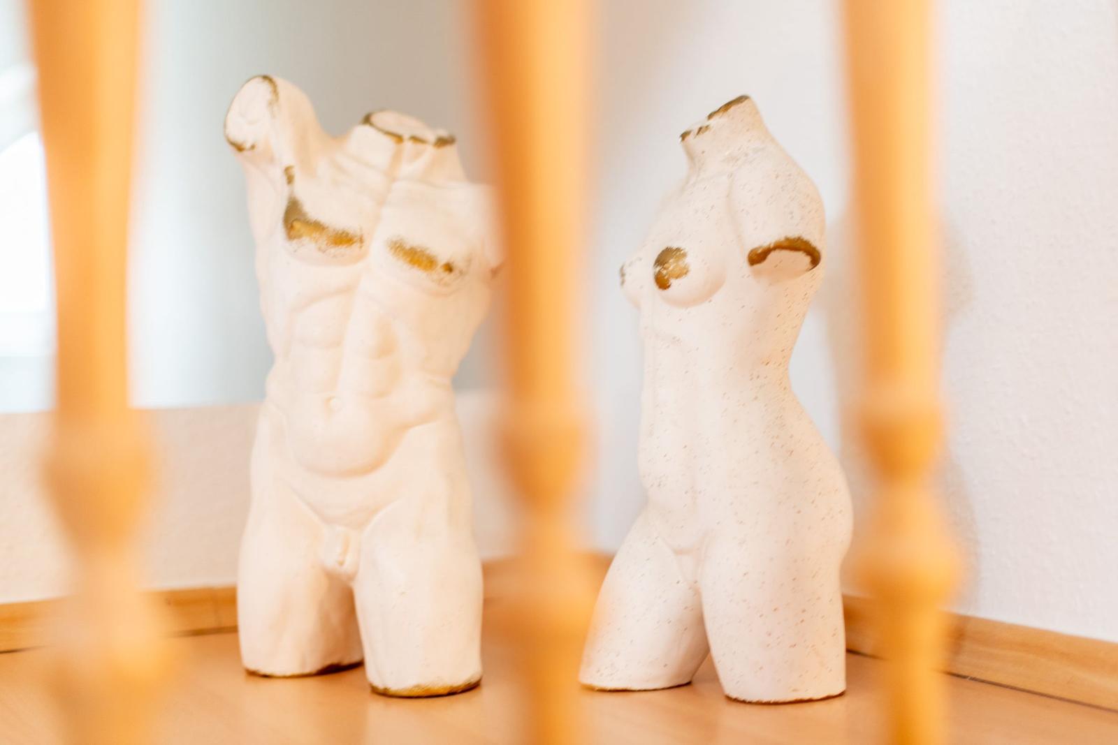 Zwei weiße Skulpturen stehen auf einem Holztisch. Eine zeigt einen männlichen Torso, die andere weiblich.