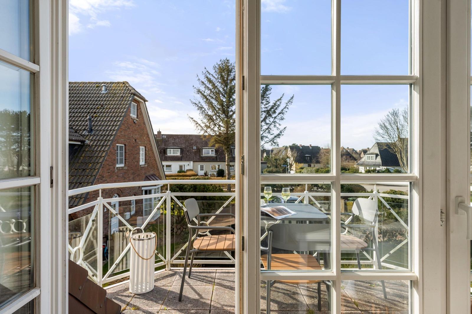 Balkon mit Tisch und Stühlen. Blick auf Häuser und Bäume. Helle Fensteröffnung.