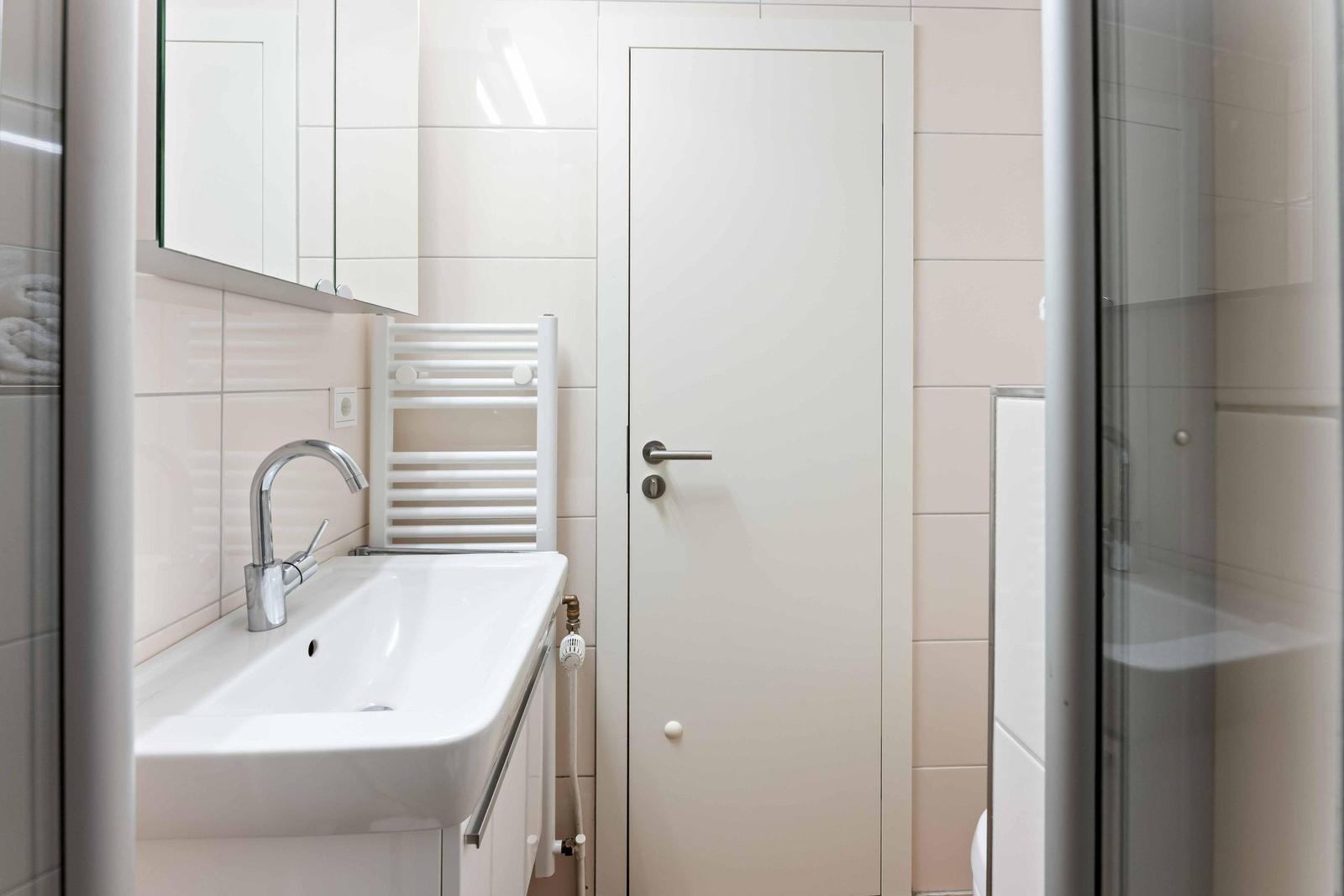Badezimmer mit Waschbecken, Spiegel und Dusche. Weiße Fliesen und Tür sichtbar.