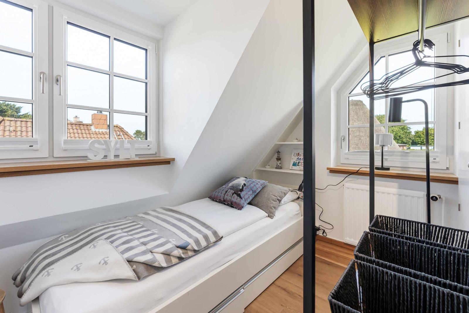 Dachgeschosszimmer mit Bett, Fenster und Schrank. Sicht auf Dächer und Grün.