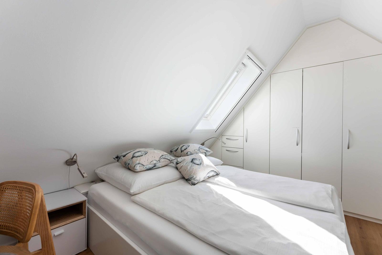 Schlafzimmer mit Dachschrägen, weißem Bett und Kleiderschrank. Fenster mit Blick nach oben.