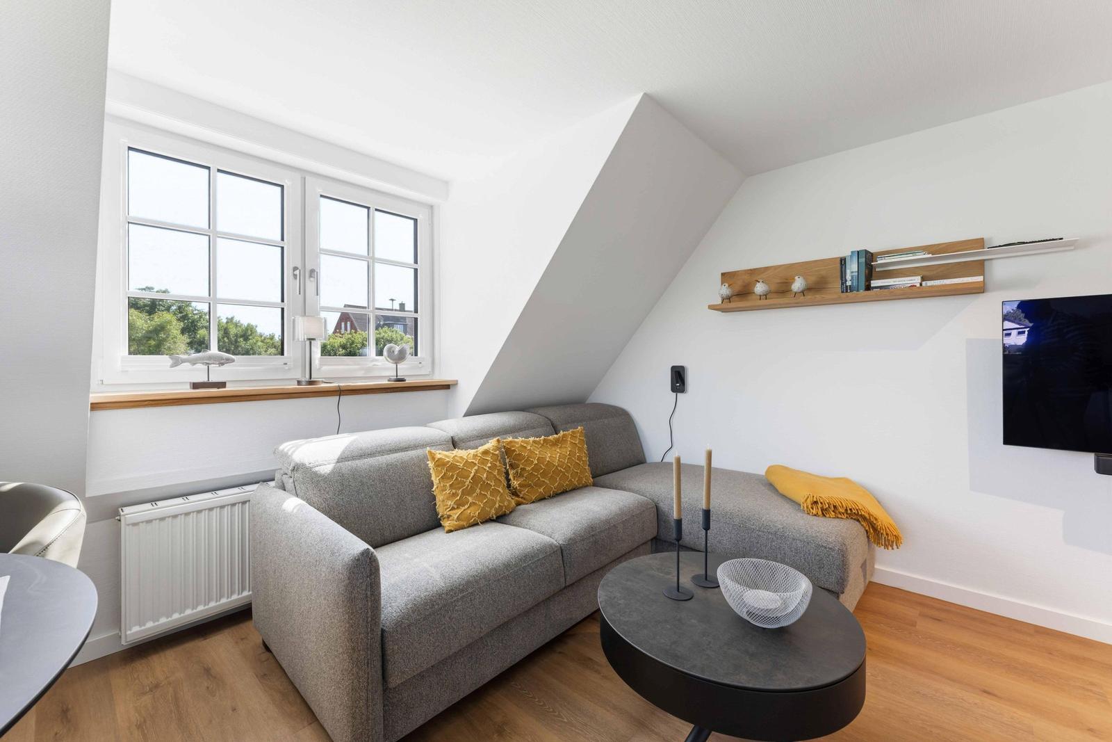 Wohnzimmer mit grauem Sofa, gelben Kissen und Holzboden. An der Wand hängt ein Fernseher.