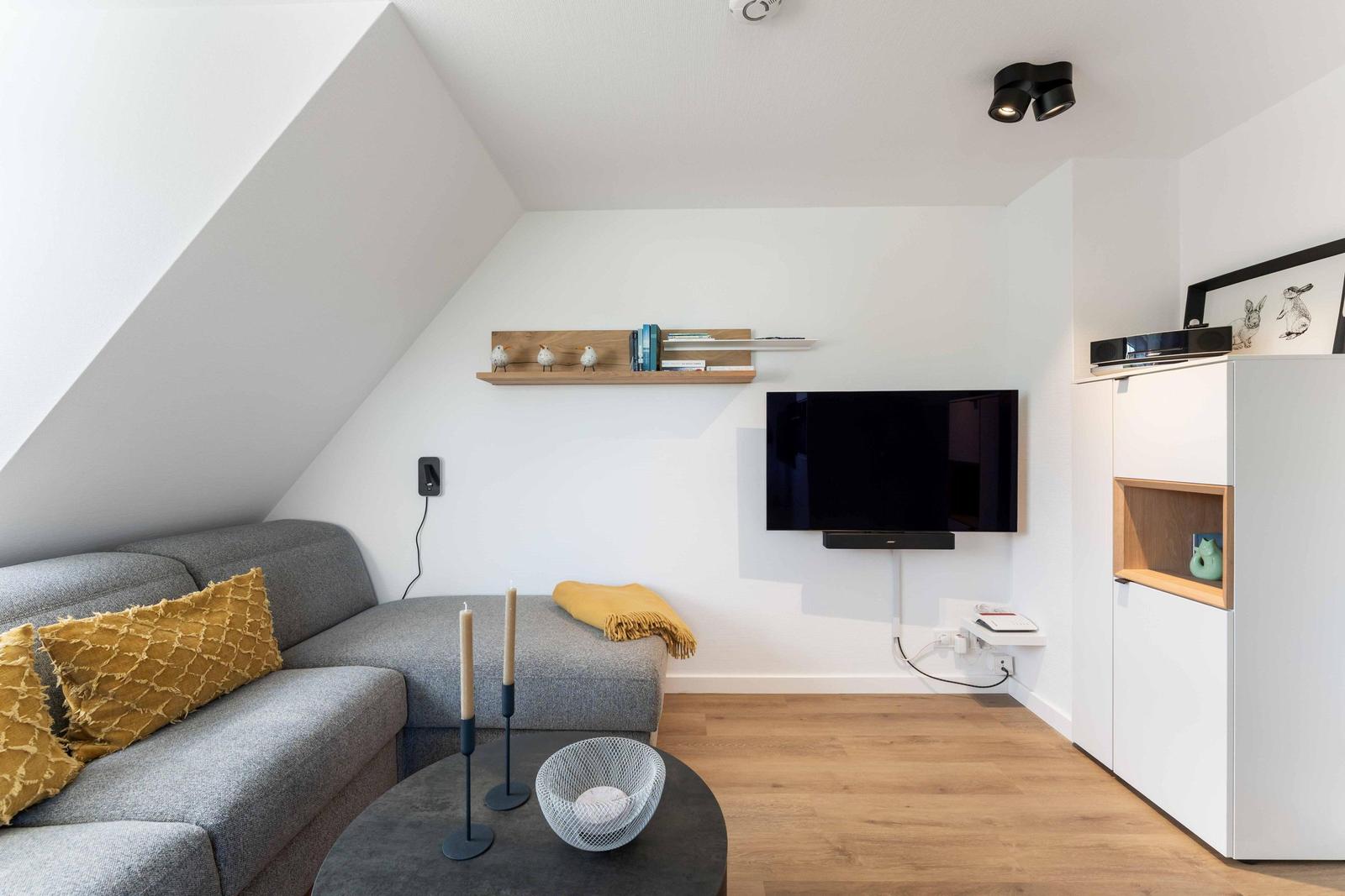Wohnzimmer mit grauem Sofa, Fernseher und Holzboden unter schrägem Dach