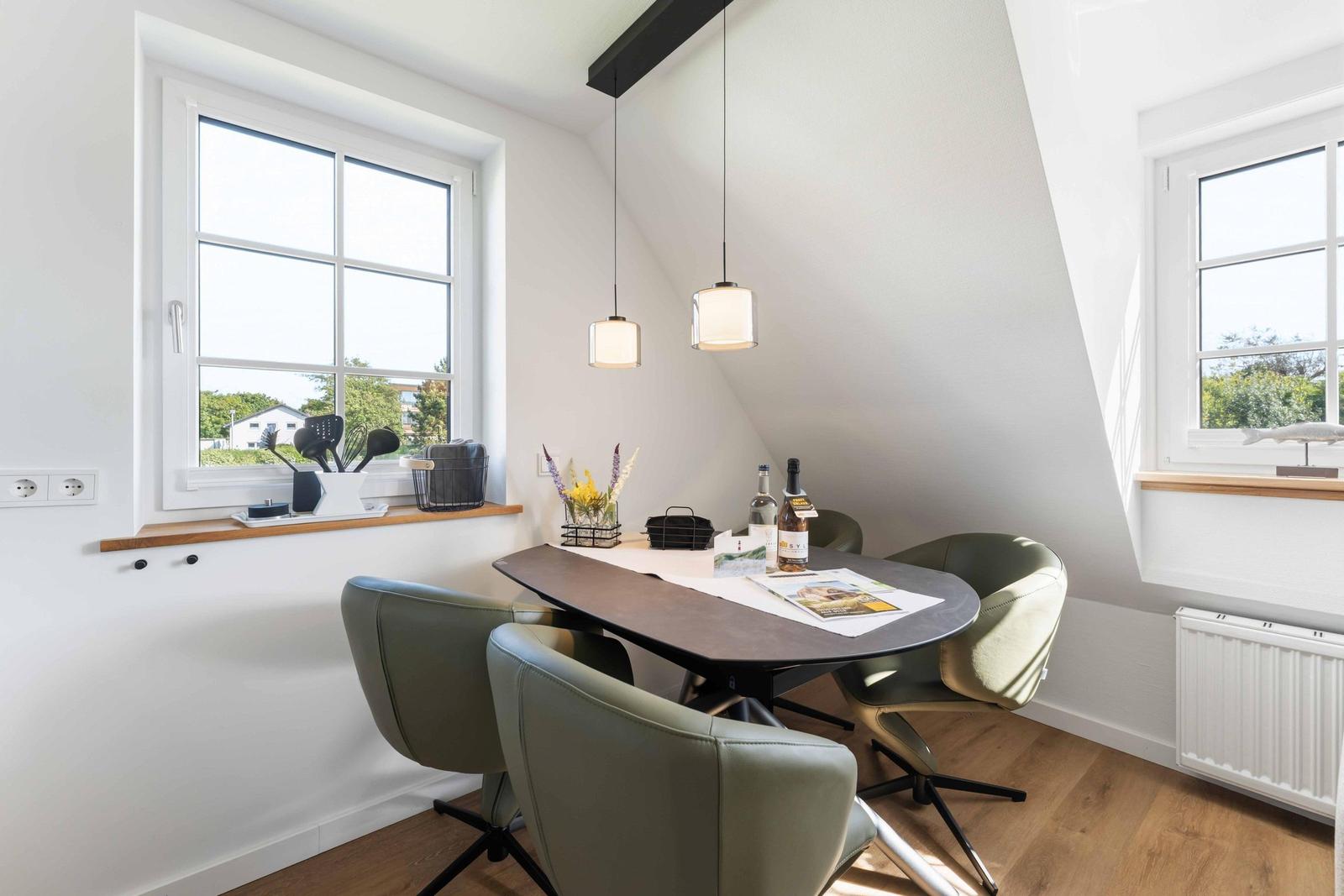 Essbereich mit Tisch, Stühlen und Fenstern. Decke mit Balken. Helle Wände und Holzboden.